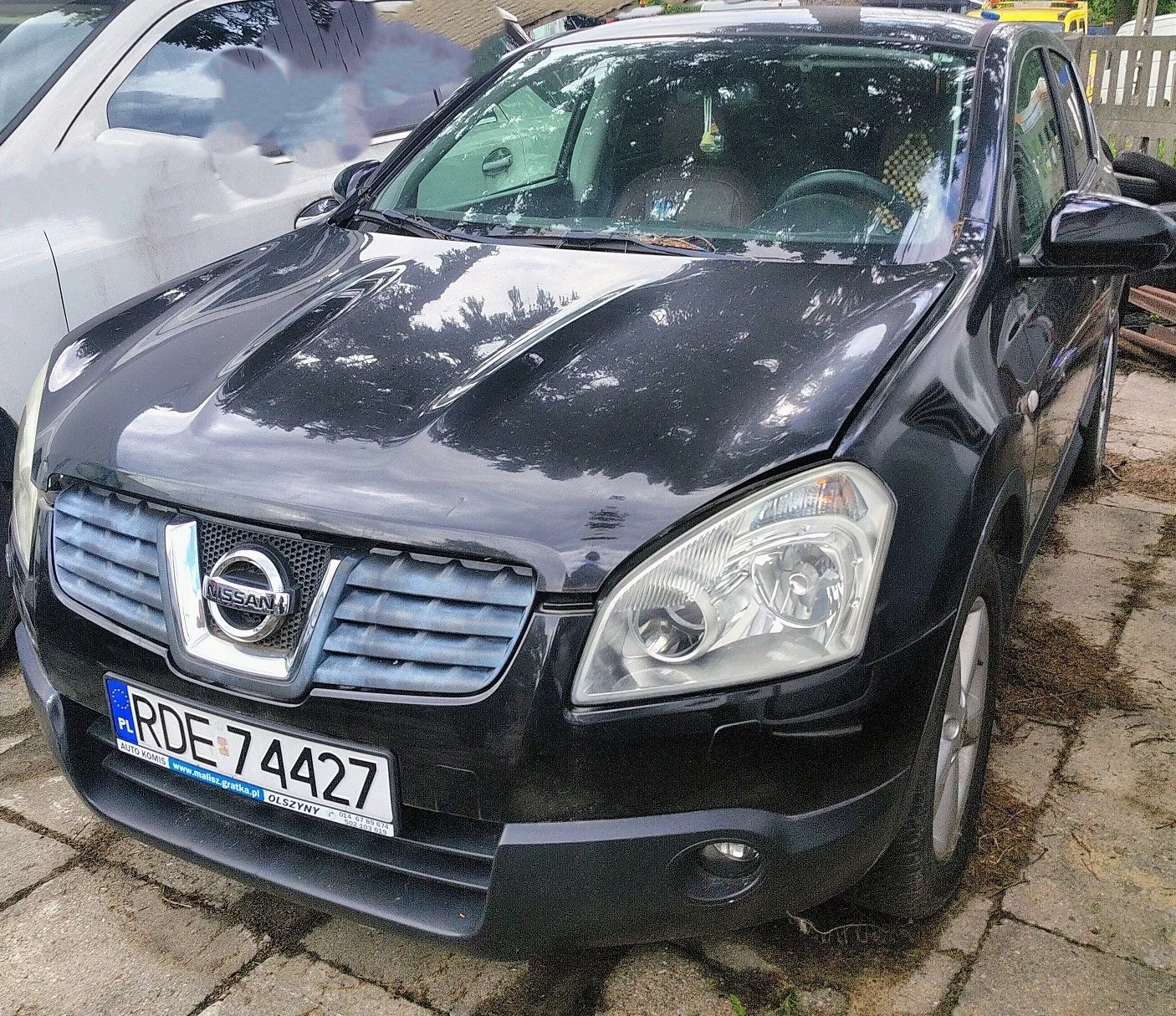 Czarny samochód marki Nissan zaparkowany na kostce brukowej. Widoczna jest przednia część pojazdu z tablicą rejestracyjną o numerze RDE 74427. Na tablicy znajduje się również napis 'AUTO-KOMIS www.multicar-gratka.pl OLSZYNY'. W tle widoczny jest fragment białego samochodu.