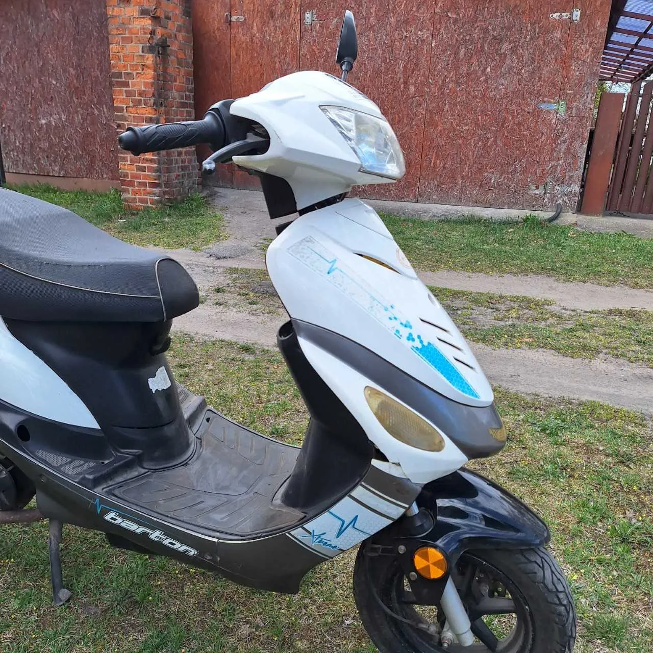 Biało-czarny skuter marki barton z niebieskimi akcentami, zaparkowany na trawie. Widoczne są czarne siedzenie, kierownica z czarnym uchwytem, przednia maska z reflektorem i pomarańczowym kierunkowskazem. Na boku skutera znajduje się napis 'barton' oraz niebieskie grafiki. W tle ceglana ściana, brązowe drzwi garażowe i trawa.