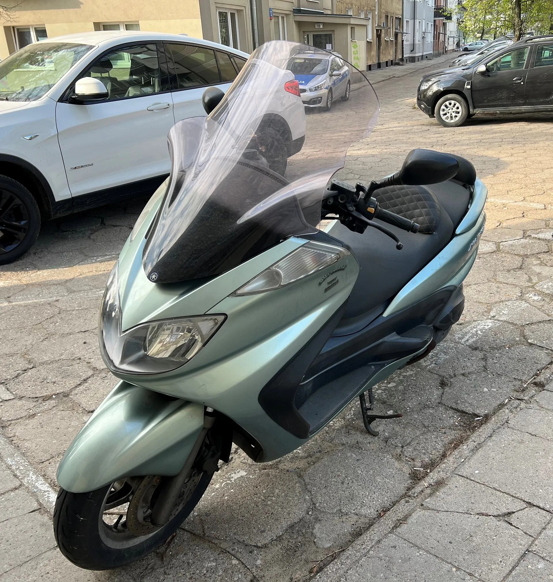 Zielony skuter marki Yamaha zaparkowany na brukowanej ulicy. W tle widoczne są samochody: biały SUV po lewej stronie, srebrny samochód i czarny SUV po prawej. Budynki mieszkalne z oknami i drzwiami w tle. Ulica wyłożona kamiennymi płytami, chodnik po prawej stronie.