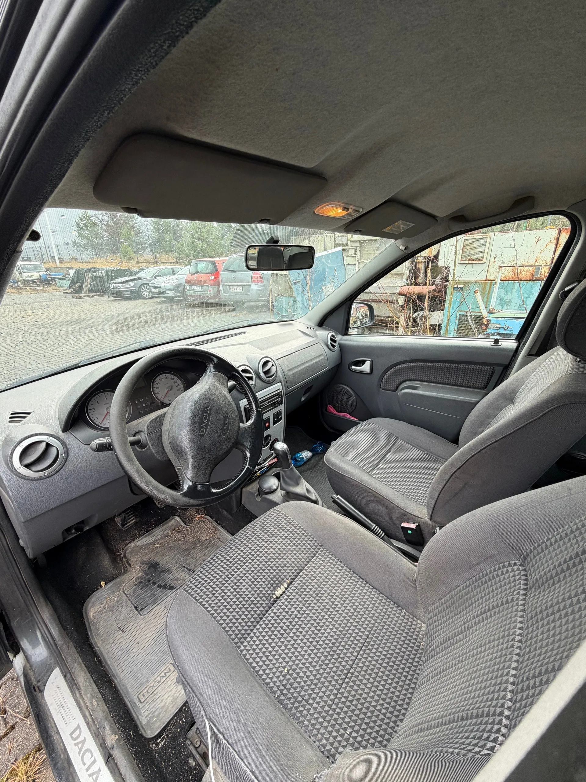 Wnętrze samochodu marki Dacia z widoczną kierownicą z logo Dacia, deską rozdzielczą z dwoma wskaźnikami, szarymi siedzeniami w geometryczny wzór, dźwignią zmiany biegów i podłogowymi matami z napisem Dacia. Przez przednią szybę widoczny parking z innymi pojazdami oraz budynkami w tle. Samochód zaparkowany na otwartym placu.