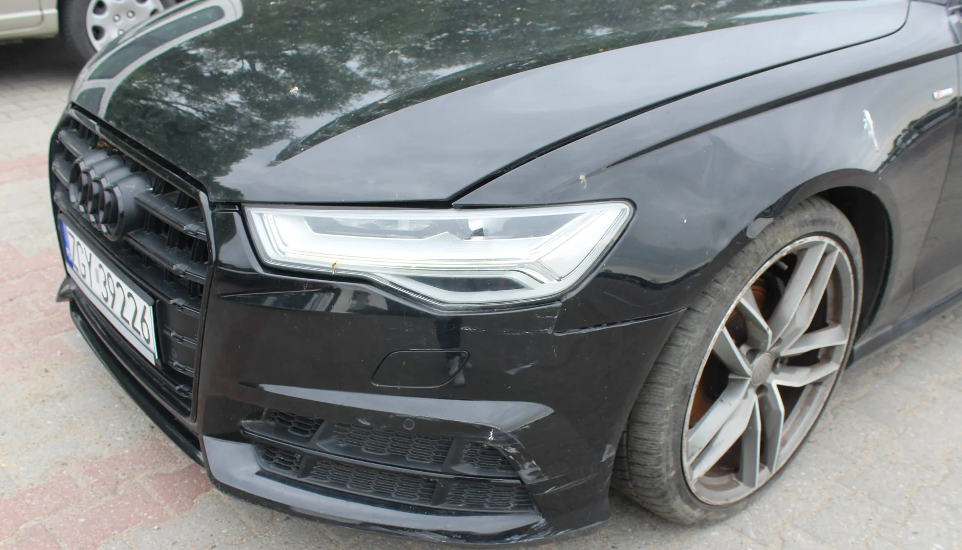 Zbliżenie na przód czarnego samochodu marki Audi. Widoczny jest lewy reflektor, fragment grilla z logo marki oraz lewe przednie koło z felgą aluminiową. Na zderzaku widoczne są zarysowania i uszkodzenia lakieru. Fragment tablicy rejestracyjnej zawiera tekst 'GY 8922K'.
