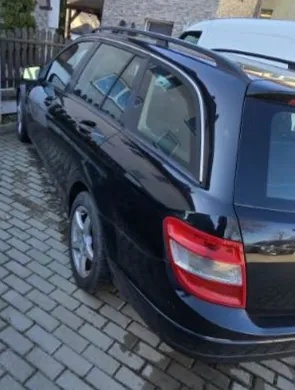 Czarny samochód typu kombi zaparkowany na brukowanej powierzchni. Widoczne są tylna część pojazdu, czerwone światła oraz fragmenty boku. W tle drewniane ogrodzenie, kamienica oraz częściowo widoczny biały samochód. Brak czytelnego tekstu na obrazie.