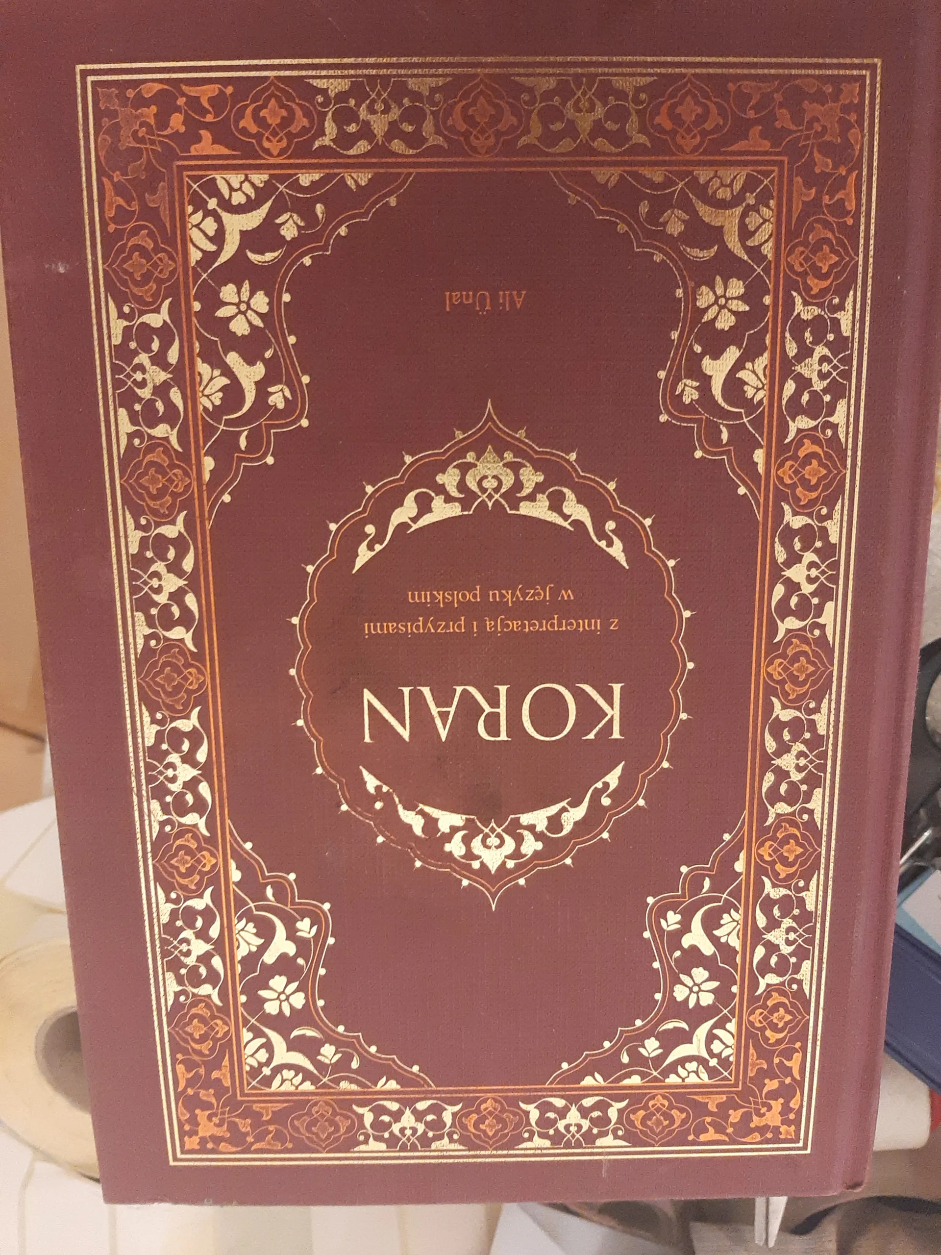 Czerwona okładka książki z złotymi i pomarańczowymi ornamentami. W centralnej części napis 'KORAN' oraz poniżej 'z interpretacją i przypisami w języku polskim' i 'Ali Ünal'. Okładka otoczona ozdobnym, geometryczno-kwiatowym wzorem. W tle widoczne są rozmyte przedmioty.