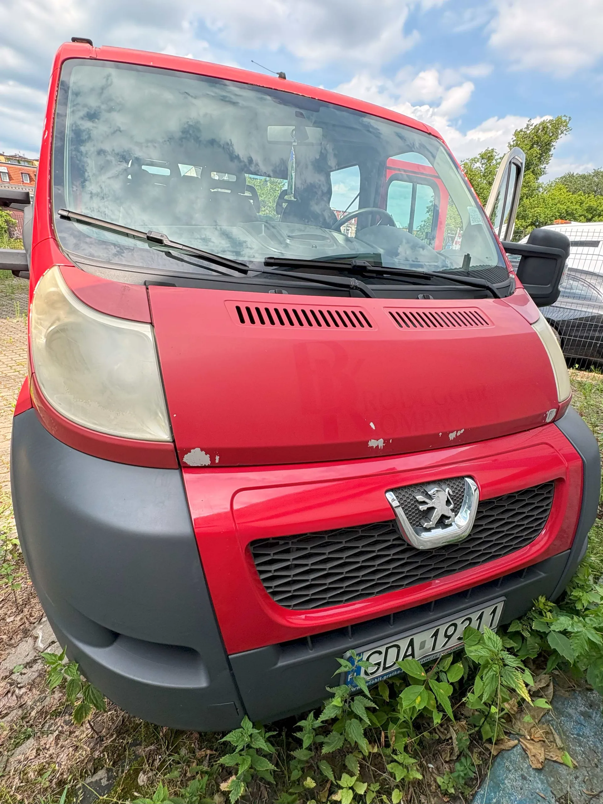 Zbliżenie na przód czerwonego vana marki Peugeot. Pojazd ma szary zderzak, zmatowione reflektory oraz widoczne odpryski lakieru na masce. Na zderzaku znajduje się tablica rejestracyjna o numerze SDA 19311. Samochód stoi na trawie pod zachmurzonym niebem.