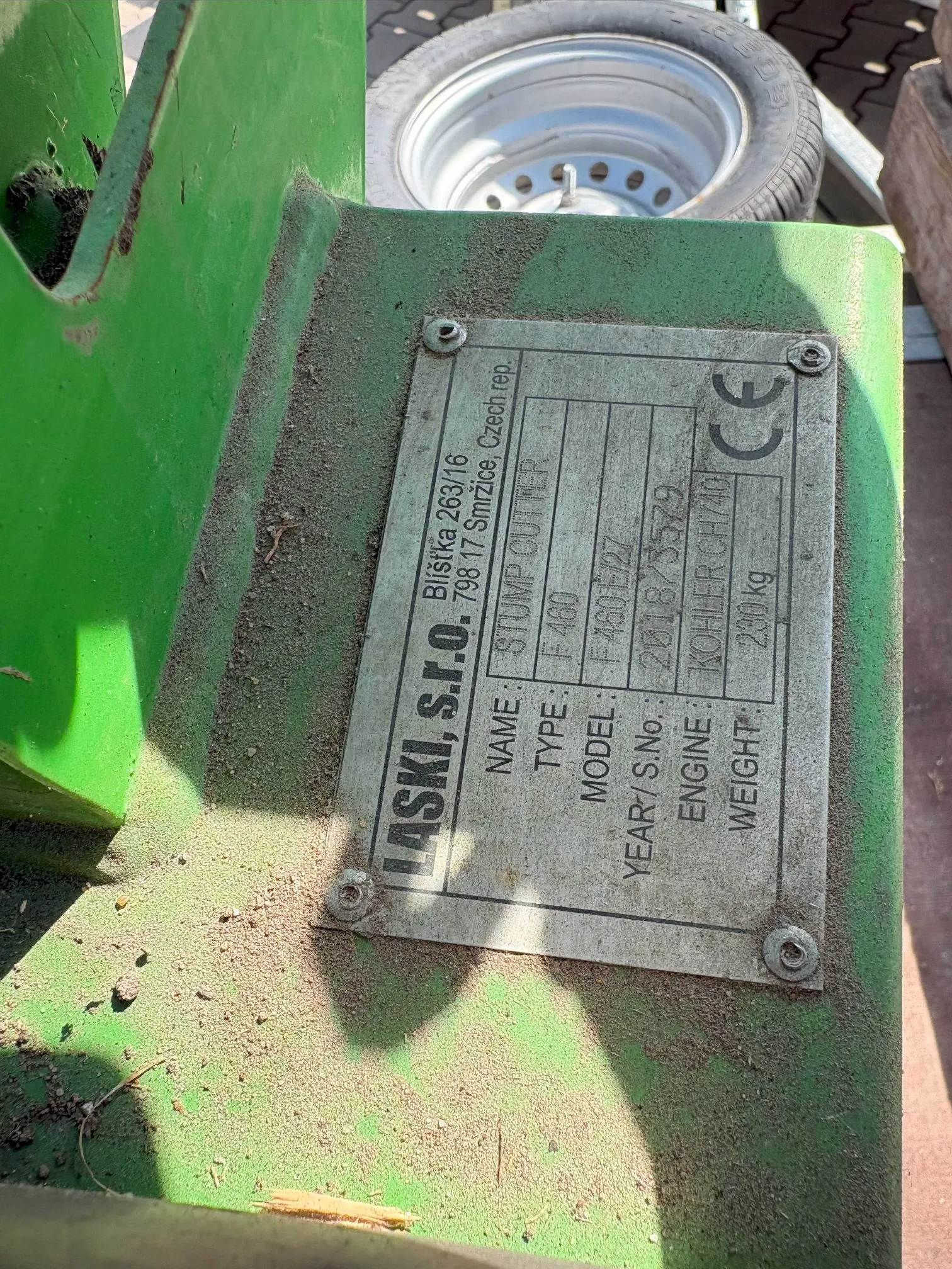 Zbliżenie na metalową tabliczkę znamionową przymocowaną do zielonej, metalowej konstrukcji maszyny. Tabliczka zawiera następujące dane: producent LASKI, s.r.o., adres Blištka 263/16, 798 17 Smřizice, Czech rep. Dane techniczne: NAME: STUMP CUTTER, TYPE: IF 480, MODEL: F 460 E/27, YEAR/S.No: 2018/3529, ENGINE: KOHLER CH740, WEIGHT: 230 kg. W prawym górnym rogu znajduje się znak CE. W tle widoczny jest fragment koła z oponą.