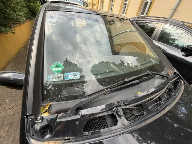 Zbliżenie na przednią szybę i maskę czarnego samochodu. Na szybie widoczne są dwie naklejki: zielona okrągła naklejka oraz niebieska prostokątna naklejka z tekstem 'GTC NETS' oraz 'GD 90 N'. Szyba jest lekko zabrudzona i odbija chmury oraz drzewa. W dolnej części widoczne są wycieraczki oraz fragment plastikowego elementu maski z kratką wentylacyjną. W tle widać fragment innego czarnego samochodu oraz żółtą ścianę budynku.