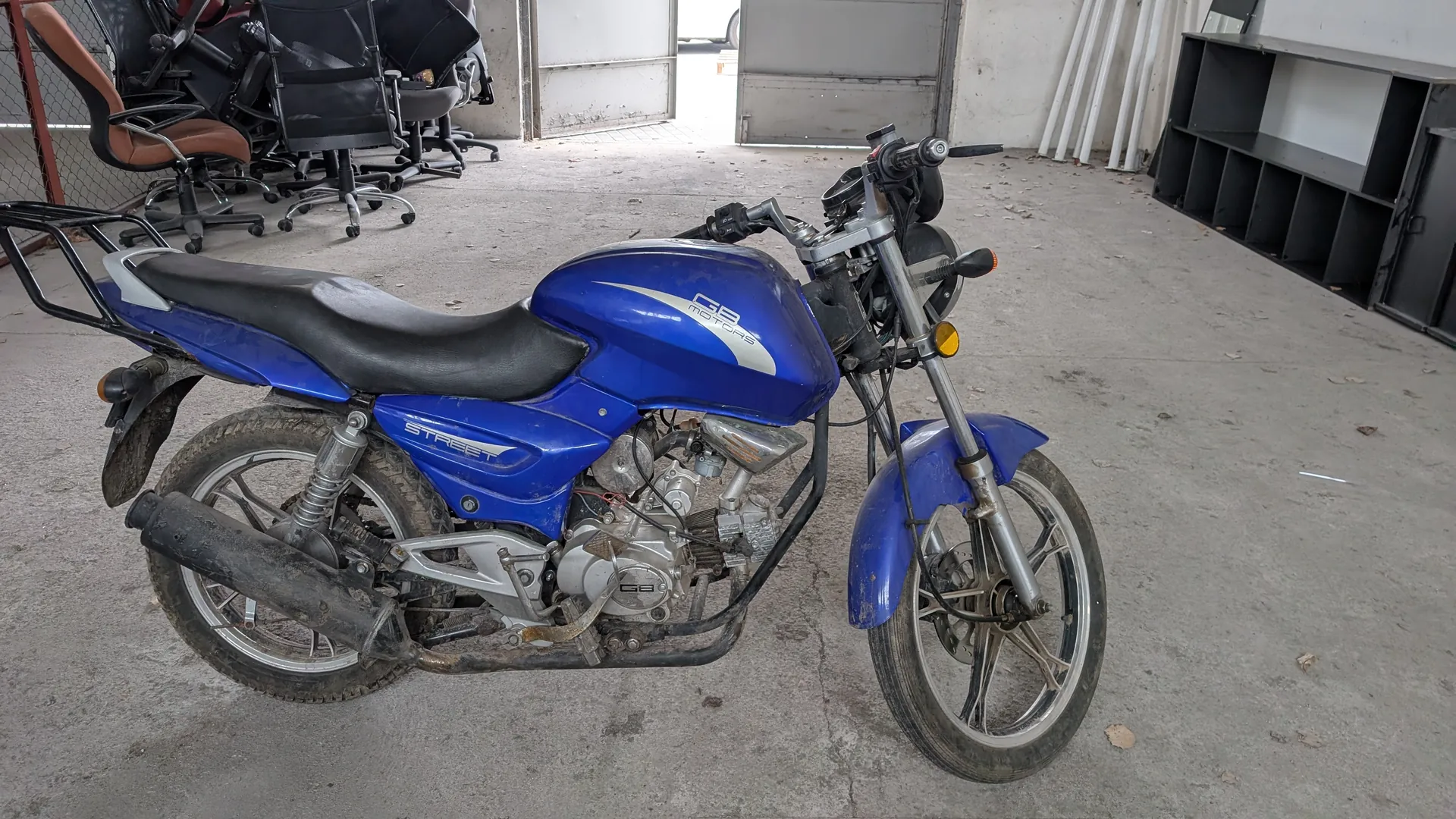 Niebieski motocykl marki GB Motors z napisem 'Street' na boku, czarnym siedzeniem i tylnym bagażnikiem. Widoczny silnik, wydech oraz koła z brudnymi oponami. W tle stos czarnych i brązowych krzeseł biurowych, betonowa podłoga z rozsypanymi liśćmi, czarna półka oraz białe rury oparte o ścianę. Przestrzeń przypomina garaż lub magazyn z otwartymi drzwiami w tle.