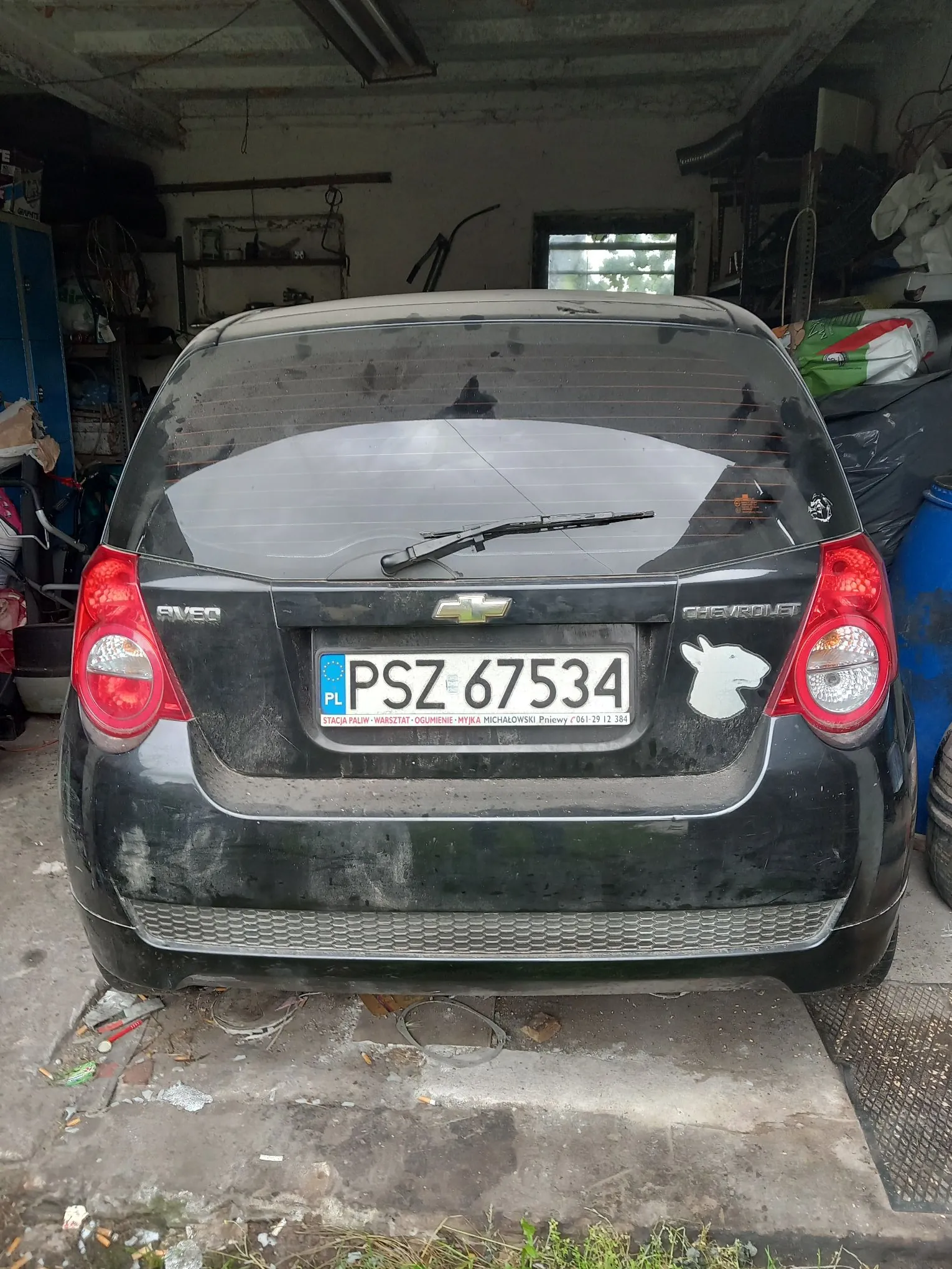 Tylny widok czarnego samochodu marki Chevrolet Aveo. Na klapie bagażnika znajduje się tablica rejestracyjna o numerze PSZ 67534 z oznaczeniem PL. Samochód jest zakurzony i znajduje się w garażu lub warsztacie.