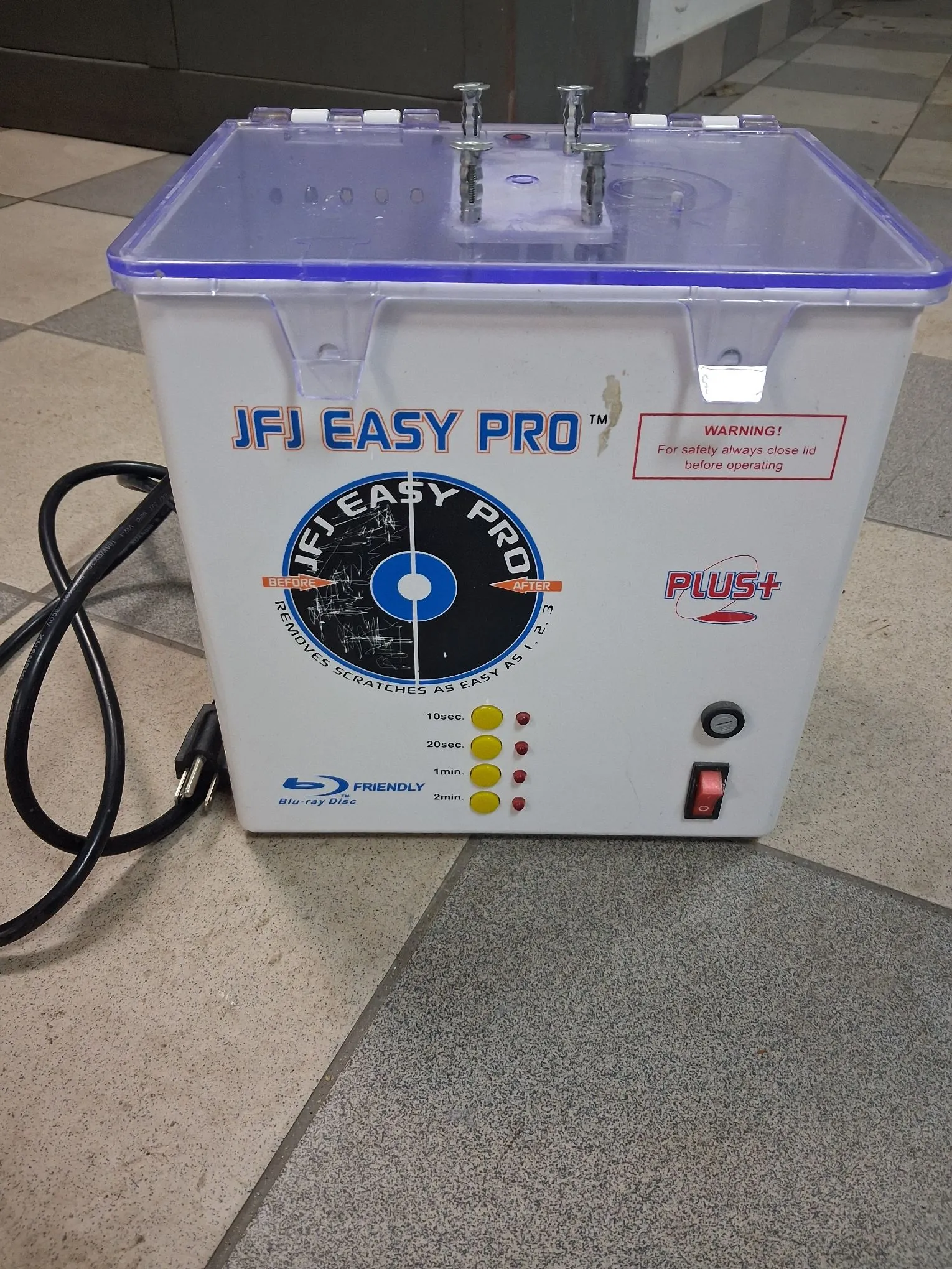 Białe urządzenie do renowacji płyt CD/DVD/Blu-ray marki JFJ EASY PRO. Urządzenie ma przezroczystą, fioletową pokrywę z uchwytami na górze. Na przednim panelu znajduje się logo 'JFJ EASY PRO', ostrzeżenie 'WARNING! For safety always close lid before operating', grafika przedstawiająca płytę z napisem 'REMOVES SCRATCHES AS EASY AS 1-2-3' oraz sekcja z czterema żółtymi przyciskami czasowymi: 10sec, 20sec, 1min, 2min. Po prawej stronie znajduje się czerwony przełącznik zasilania, a po lewej czarny kabel z wtyczką. Na dole widnieje napis 'Blu-ray Disc FRIENDLY'.