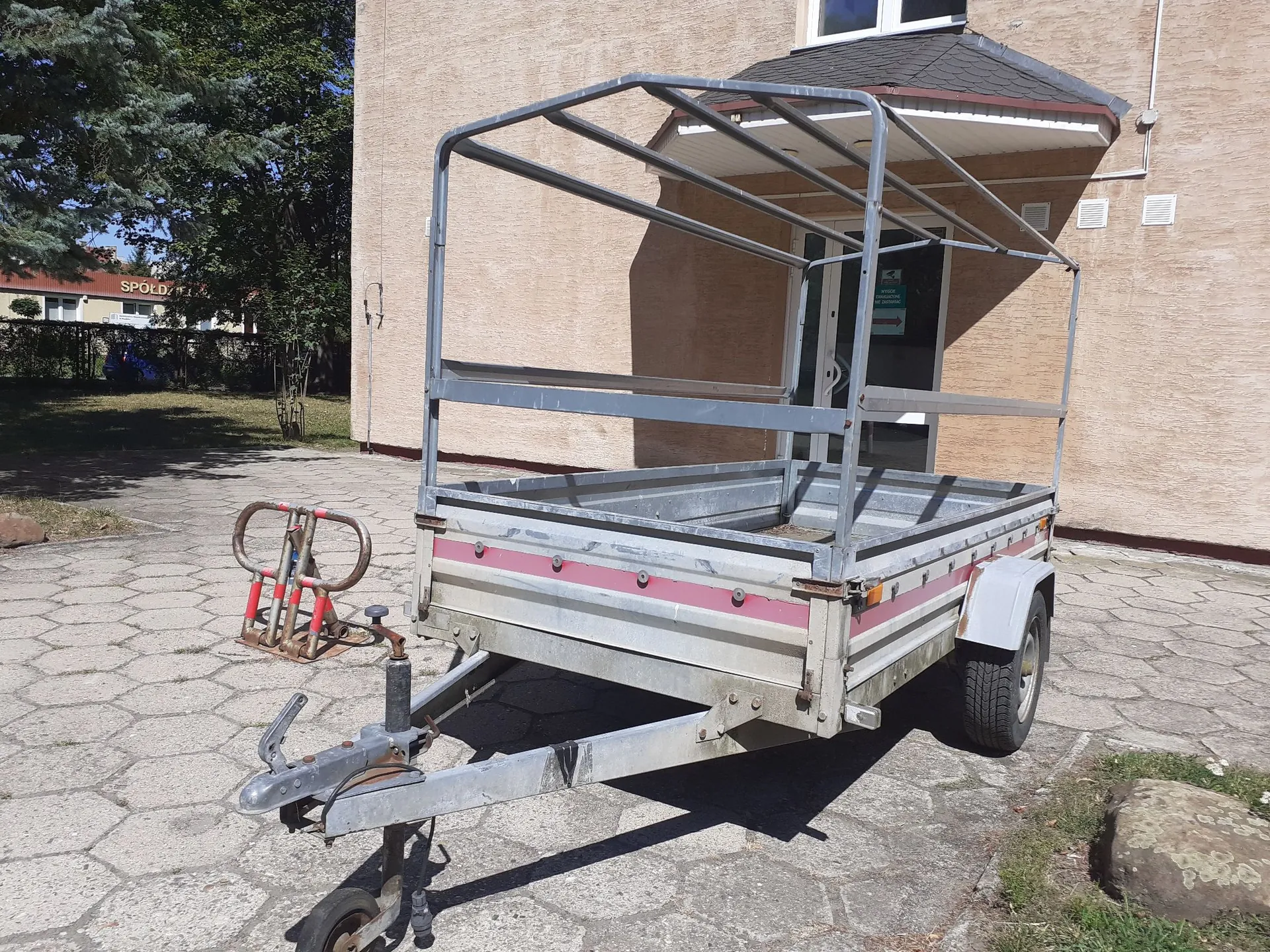 Srebrna przyczepa transportowa z metalową konstrukcją stelaża nad paką, zaparkowana na betonowym placu przed beżowym budynkiem. Przyczepa ma czerwony pas dekoracyjny na boku i jest wyposażona w dyszel z kołem podporowym oraz uchwytem rowerowym z przodu.