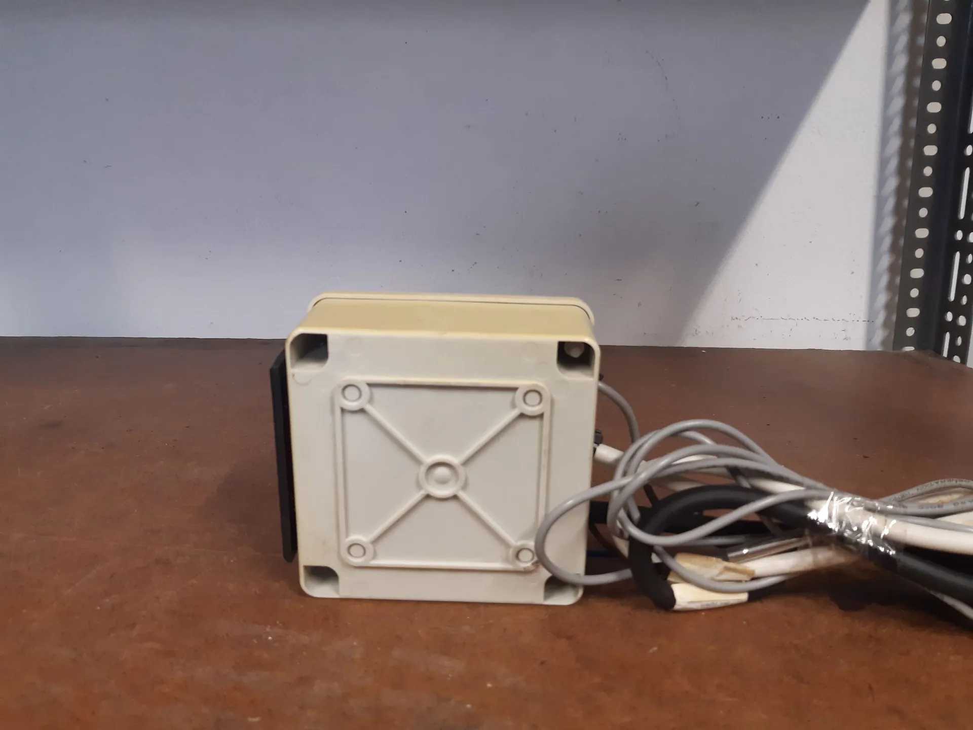 Kremowa, plastikowa obudowa urządzenia elektronicznego w kształcie sześcianu, ustawiona na brązowej powierzchni. Na przedniej ściance obudowy widoczny jest wypukły wzór w kształcie litery X z czterema śrubami w rogach. Z prawej strony urządzenia wychodzą szare i czarne przewody elektryczne.