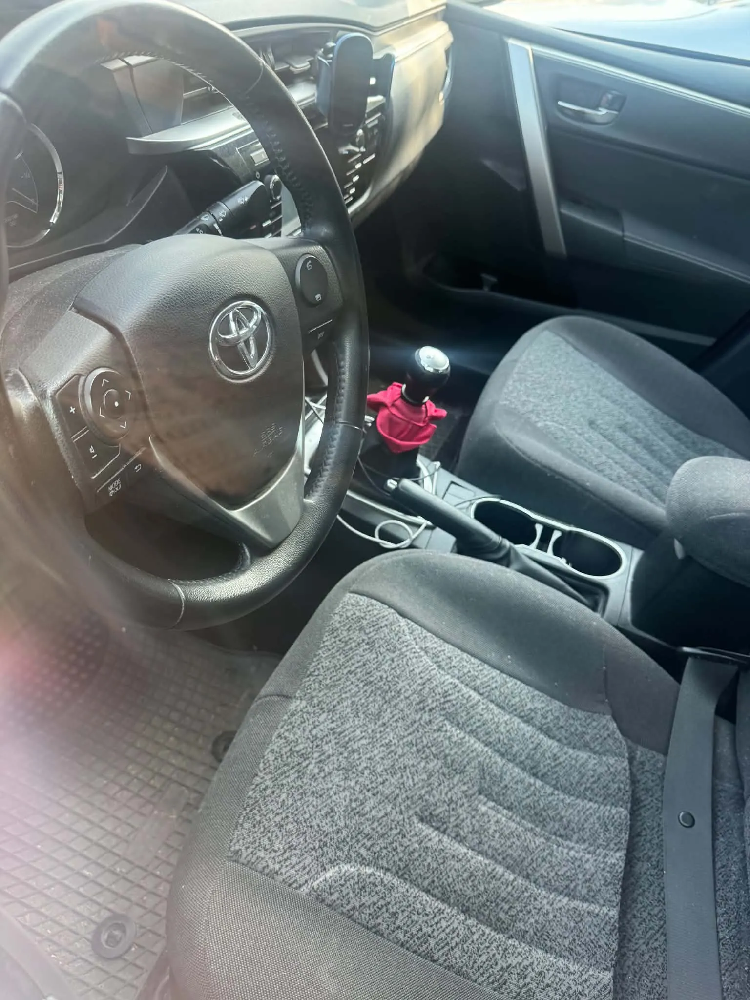 Wnętrze samochodu marki Toyota widziane z perspektywy kierowcy. Widoczna jest czarna kierownica z logo Toyoty, dwa przednie fotele z szaro-czarną tapicerką materiałową, lewarek zmiany biegów z czerwoną osłoną oraz konsola środkowa z uchwytami na kubki.