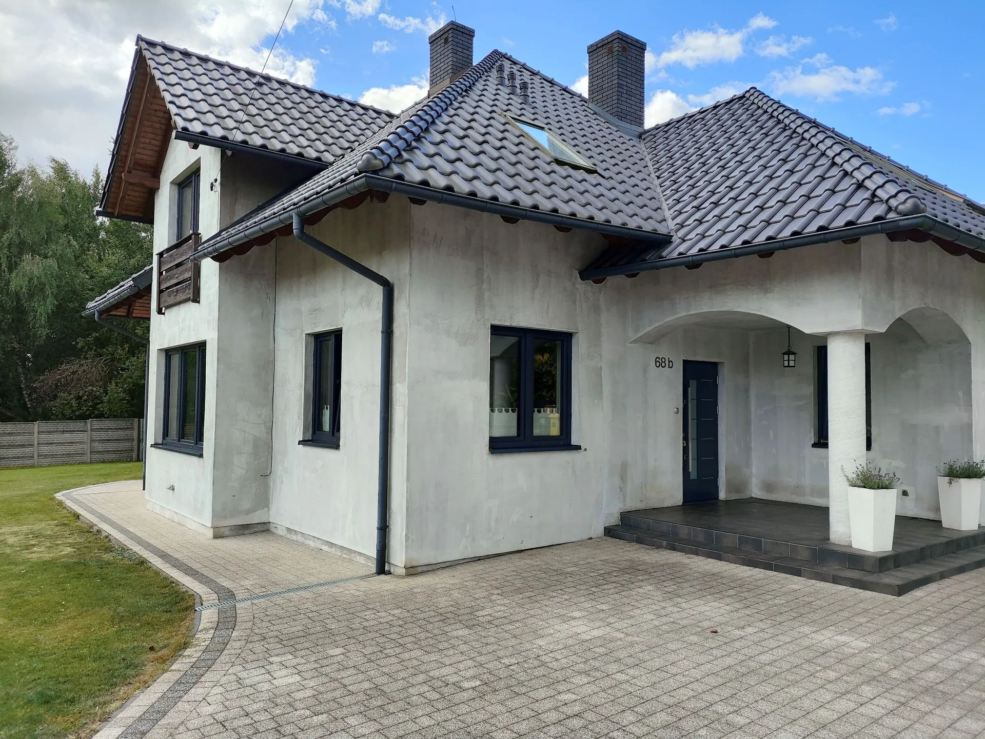 Nowoczesny, parterowy dom z poddaszem, wykończony szarym tynkiem strukturalnym. Budynek posiada ciemnoszary, spadzisty dach z dachówek, dwa kominy oraz okno połaciowe. Widoczne są ciemne ramy okienne i ciemne drzwi wejściowe z numerem domu '68 b' nad nimi. Przed wejściem znajduje się zadaszony taras z białymi kolumnami i dwiema białymi donicami z roślinami. Przed domem rozciąga się podjazd z szarej kostki brukowej, a obok niego pas zielonej trawy.