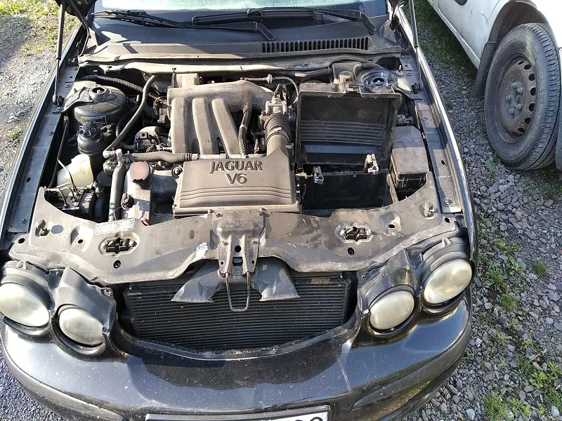 Czarny samochód Jaguar z otwartą maską, ukazujący silnik oznaczony napisem 'JAGUAR V6'. Widoczne są elementy silnika, w tym układ chłodzenia, przewody i reflektory. Samochód stoi na żwirowym podłożu z trawą, obok częściowo widoczny biały pojazd.