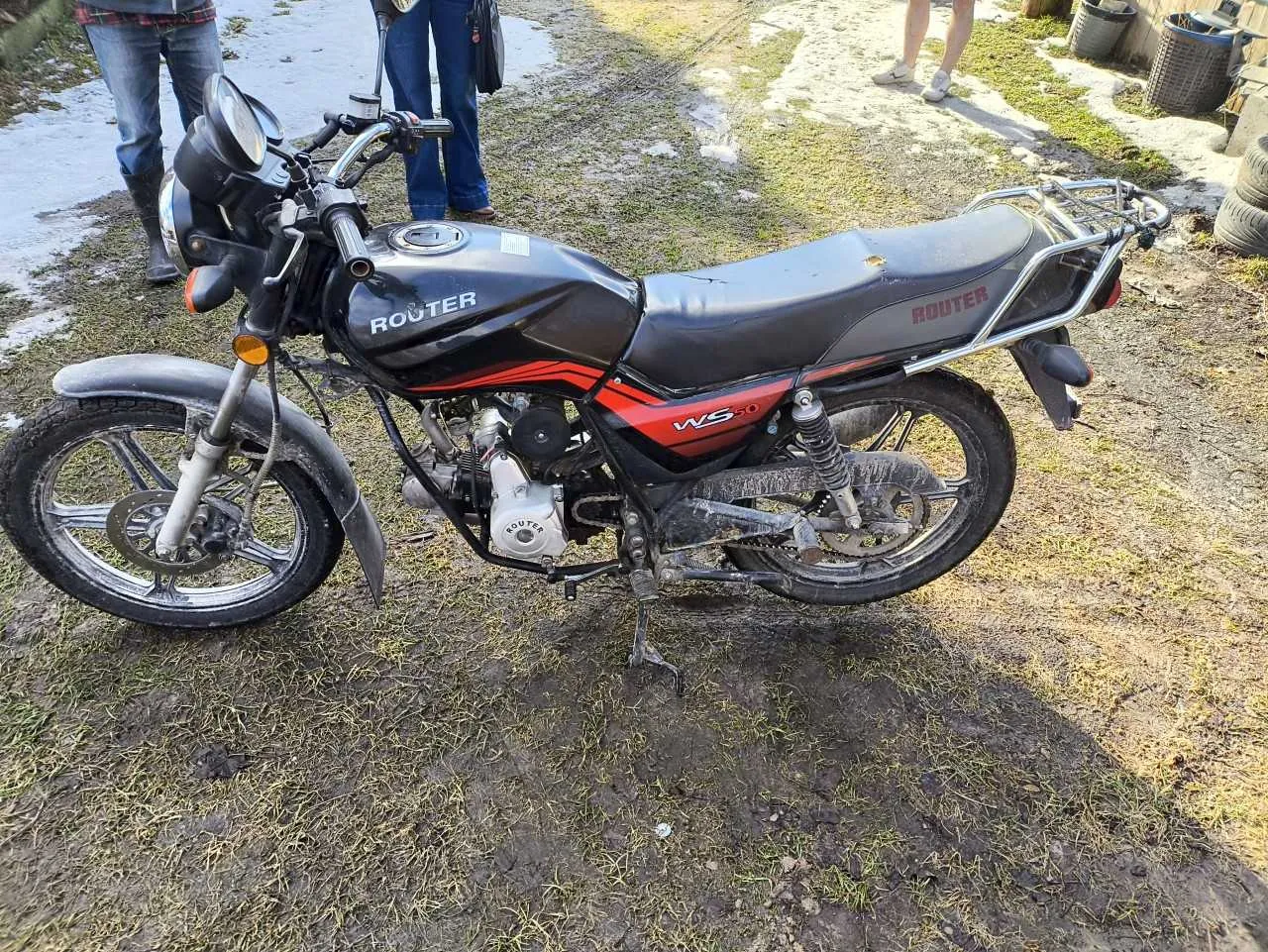 Czarno-czerwony motocykl marki Router model WS-10 zaparkowany na trawiastym i błotnistym terenie. Na zbiorniku paliwa widoczny napis 'ROUTER', na boku czerwonej części napis 'WS-10'. W tle stoją trzy osoby: jedna w dżinsach i czarnych butach, druga w dżinsach z torebką, trzecia w białych butach. Po prawej stronie widoczne są opony i metalowy pojemnik. Motocykl ma czarne siedzenie, chromowane elementy i błotniki.