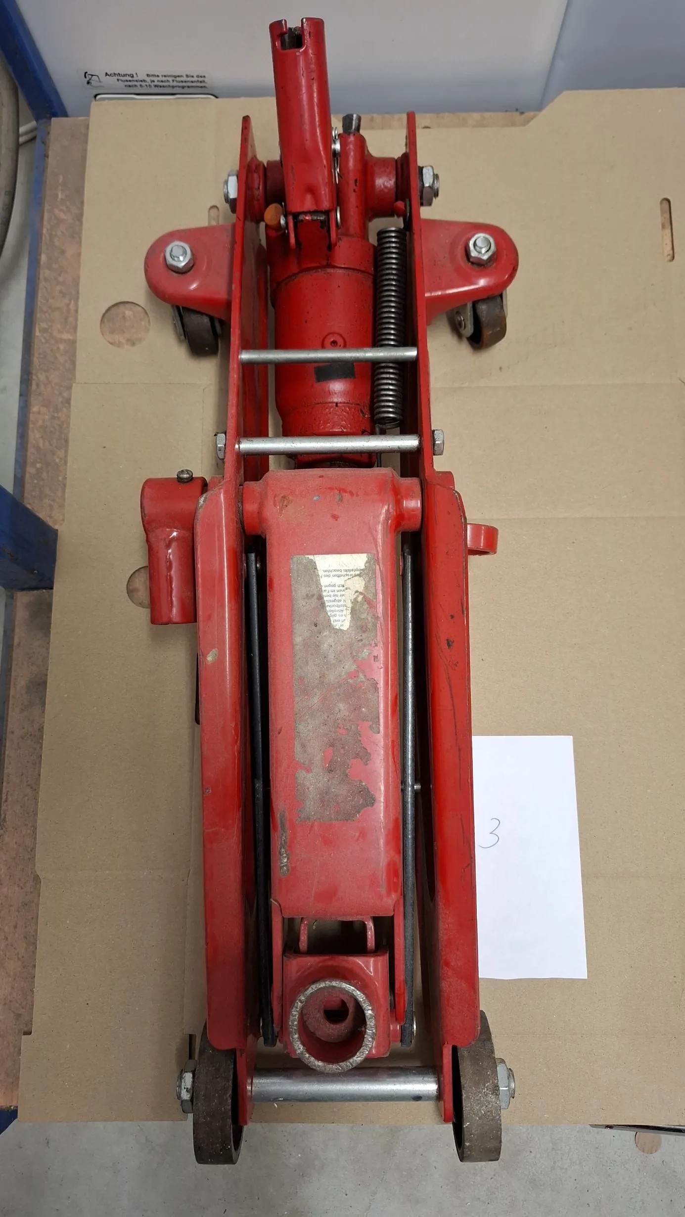 Czerwony podnośnik hydrauliczny do samochodów, ułożony pionowo na brązowym kartonie. Urządzenie posiada kółka u góry i na dole oraz srebrne elementy metalowe i sprężynę. Na boku znajduje się częściowo starte, białe pole z tekstem. Obok podnośnika leży biała kartka z napisem '3'.