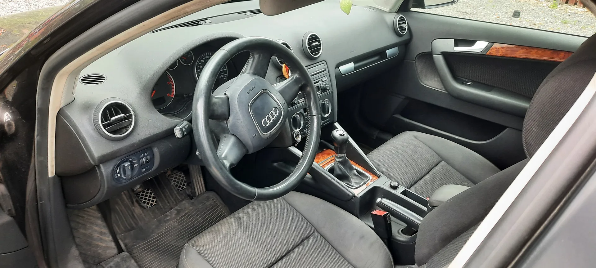 Wnętrze samochodu marki Audi z widocznym kierownicą z logo czterech pierścieni, szarą deską rozdzielczą z okrągłymi wydmuchami, wskaźnikami prędkości i obrotomierzem. W centrum konsoli znajduje się dźwignia zmiany biegów, a siedzenia są tapicerowane szarym materiałem. Drzwi posiadają wykończenie z drewnianą listwą. Podłoga pokryta jest brudnymi dywanikami. Wnętrze jest ujęte z perspektywy otwartego drzwi pasażera.