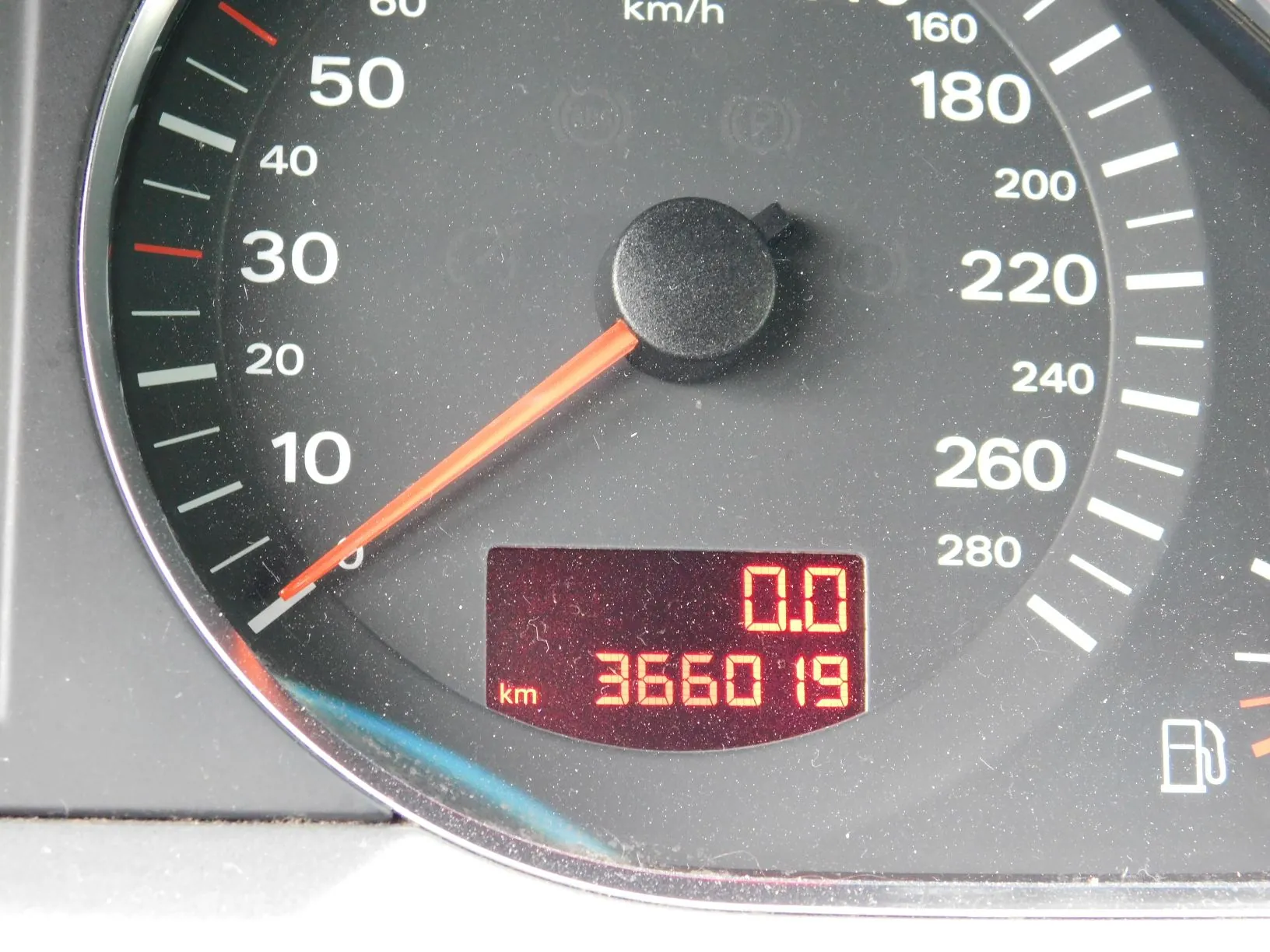 Zbliżenie na prędkościomierz w samochodzie. Tarcza z białymi cyframi od 10 do 280 km/h na czarnym tle. Pomarańczowa wskazówka wskazuje wartość bliską 0. Na dole znajduje się cyfrowy wyświetlacz z czerwonymi cyframi, pokazujący wartość 0.0 oraz przebieg 366019 km.