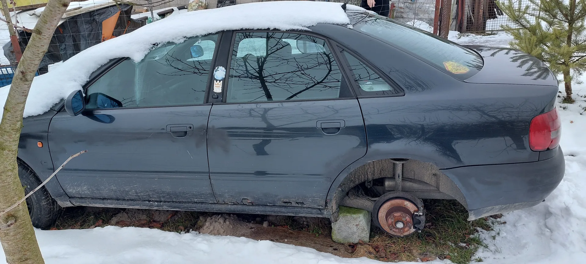 Ciemnoszary samochód osobowy typu sedan z pokrytą śniegiem dachem, zaparkowany na śnieżnej powierzchni. Brakuje tylnego prawego koła, widoczny jest zardzewiały tarcza hamulcowa i podparta betonowym blokiem osi. Na bocznych szybach naklejki, w tym z koroną i niebieskim symbolem. W tle drzewa, siatka ogrodzeniowa, budynki i śnieg na ziemi. Samochód pokryty warstwą brudu i śniegu.