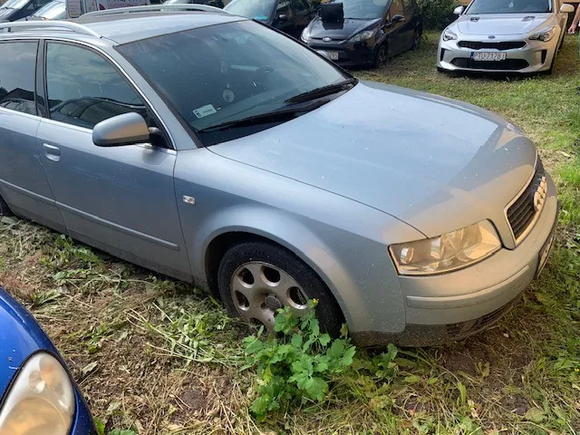 Srebrny samochód marki Audi w nadwoziu kombi, zaparkowany na trawiastym terenie. Pojazd jest widoczny z przodu i z boku, z naciskiem na przednie prawe koło, które znajduje się w niskiej trawie i roślinności. W tle widoczne są inne samochody, w tym biały pojazd z tablicą rejestracyjną RI 7128J.