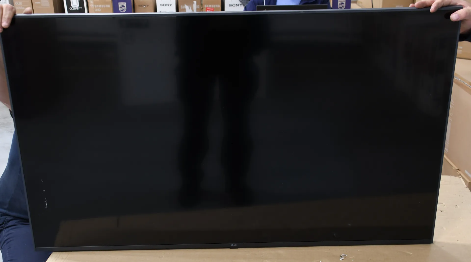 Zdjęcie przedstawia osobę trzymającą duży, wyłączony telewizor z czarnym ekranem. W tle widoczne są kartonowe opakowania sprzętu elektronicznego marek Samsung i Sony. Na dolnej ramce telewizora znajduje się małe logo LG.