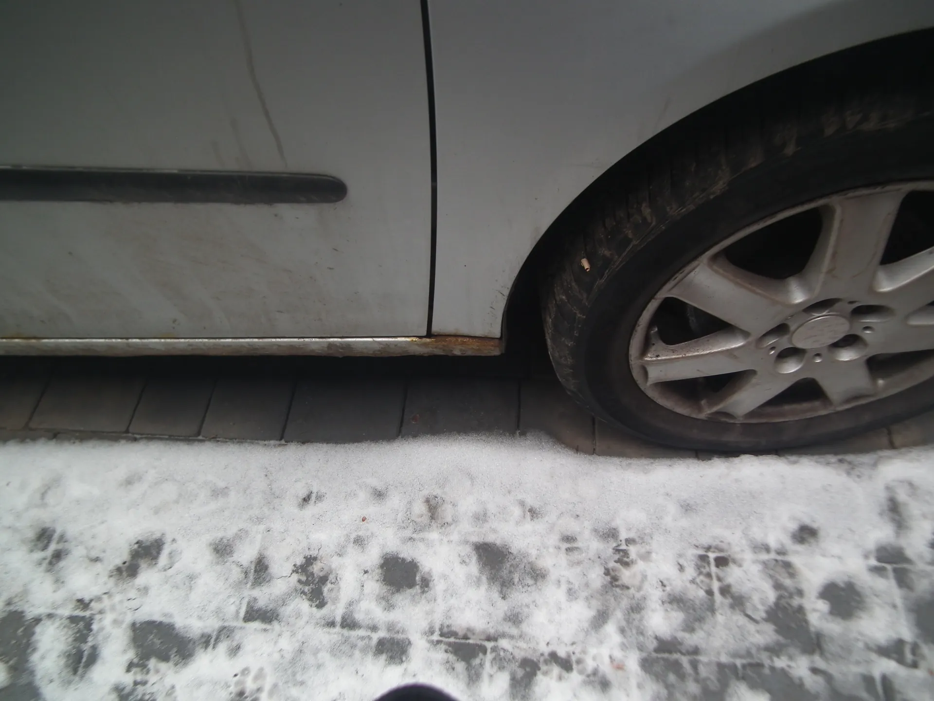Fragment białego samochodu z widocznym kołem i częścią pasa bocznego. Na dnie drzwi widoczna rdza, a na oponie brud. Pod samochodem chodnik z ciemnych płytek i warstwa śniegu z odciskami stóp. Koło ma srebrną felgę z pięcioma ramionami.