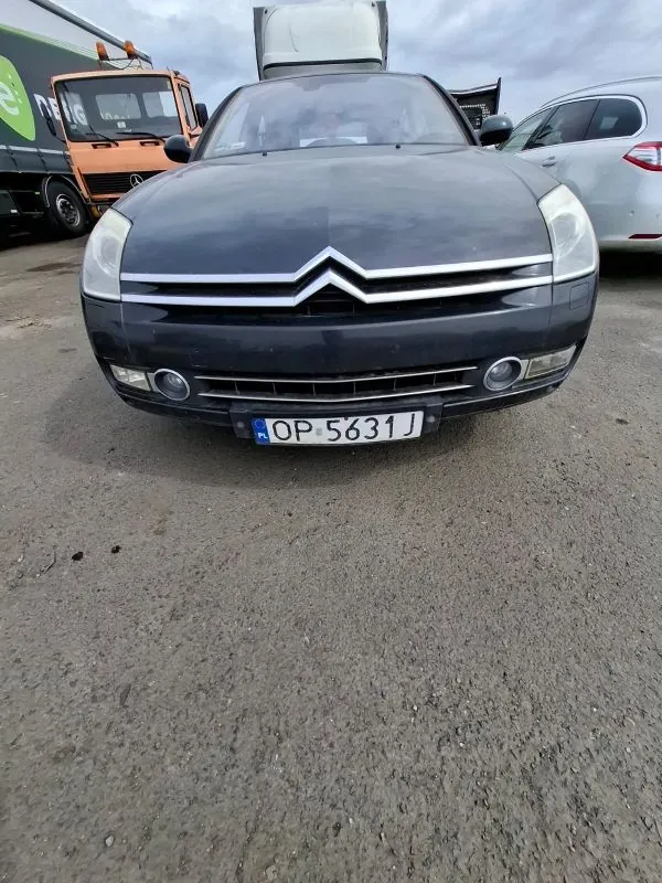 Czarny samochód marki Citroën z przodu, z widocznym logo na masce i tablicą rejestracyjną OP 5631 J. W tle po lewej stronie pomarańczowy ciężarówka z częściowo widocznym zielonym logo i napisem, po prawej srebrny samochód. Asfaltowa powierzchnia z drobnymi śladami, pochmurne niebo. Tekst na tablicy rejestracyjnej jest czytelny i ustawiony poziomo.