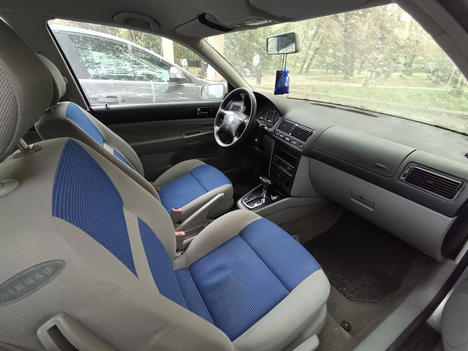 Wnętrze samochodu widziane z perspektywy tylnego siedzenia. Widoczne są dwa przednie fotele z szaro-niebieską tapicerką, deska rozdzielcza, kierownica z logo Volkswagena oraz lewarek zmiany biegów. Przez przednią szybę i boczną szybę kierowcy widoczny jest widok na zewnątrz z drzewami i innym zaparkowanym samochodem.