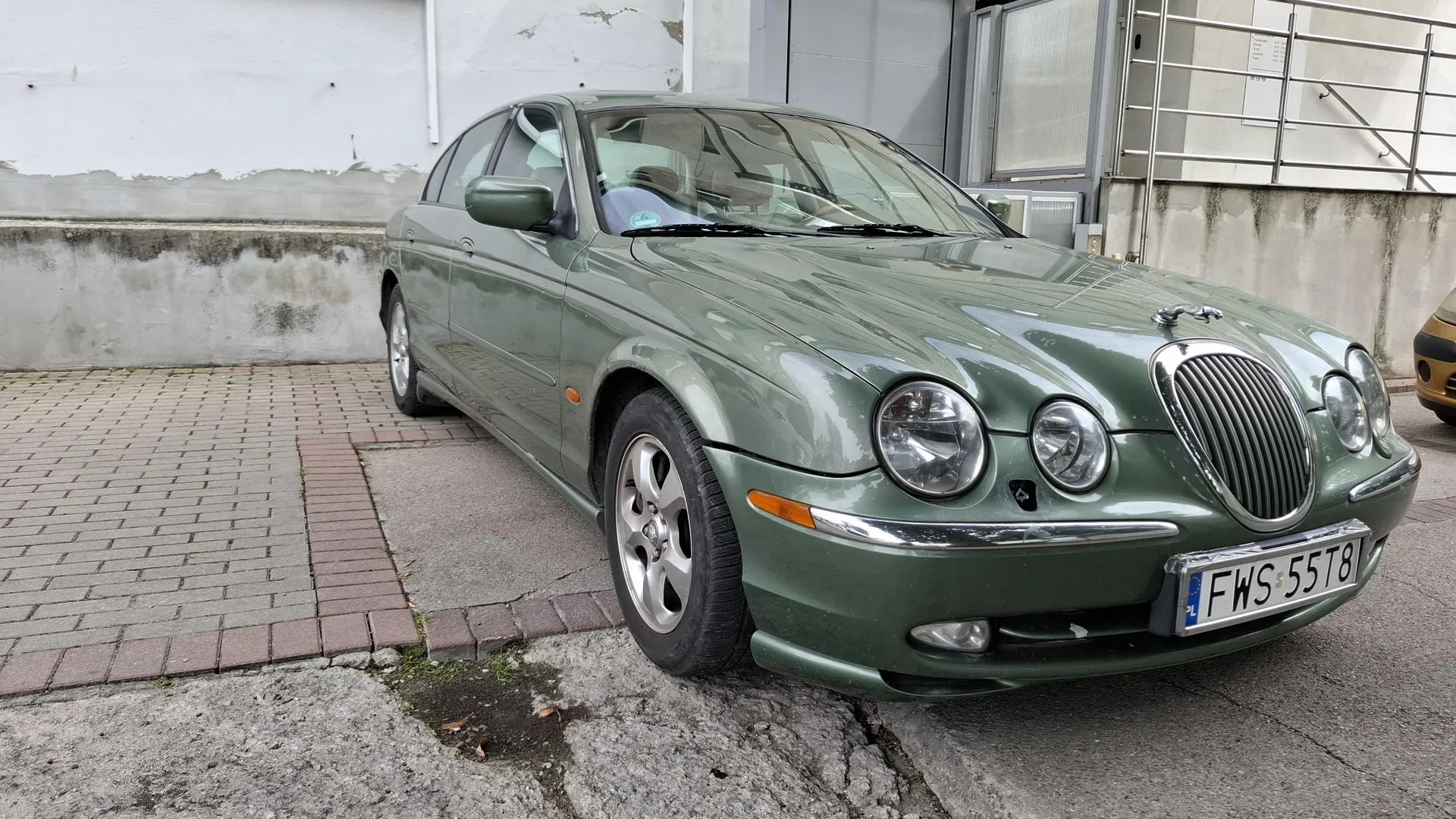 Srebrno-zielony samochód osobowy marki Jaguar zaparkowany na kostce brukowej przed jasną ścianą budynku. Pojazd jest widoczny z przodu pod kątem, z widoczną tablicą rejestracyjną o treści FWS 55T8.