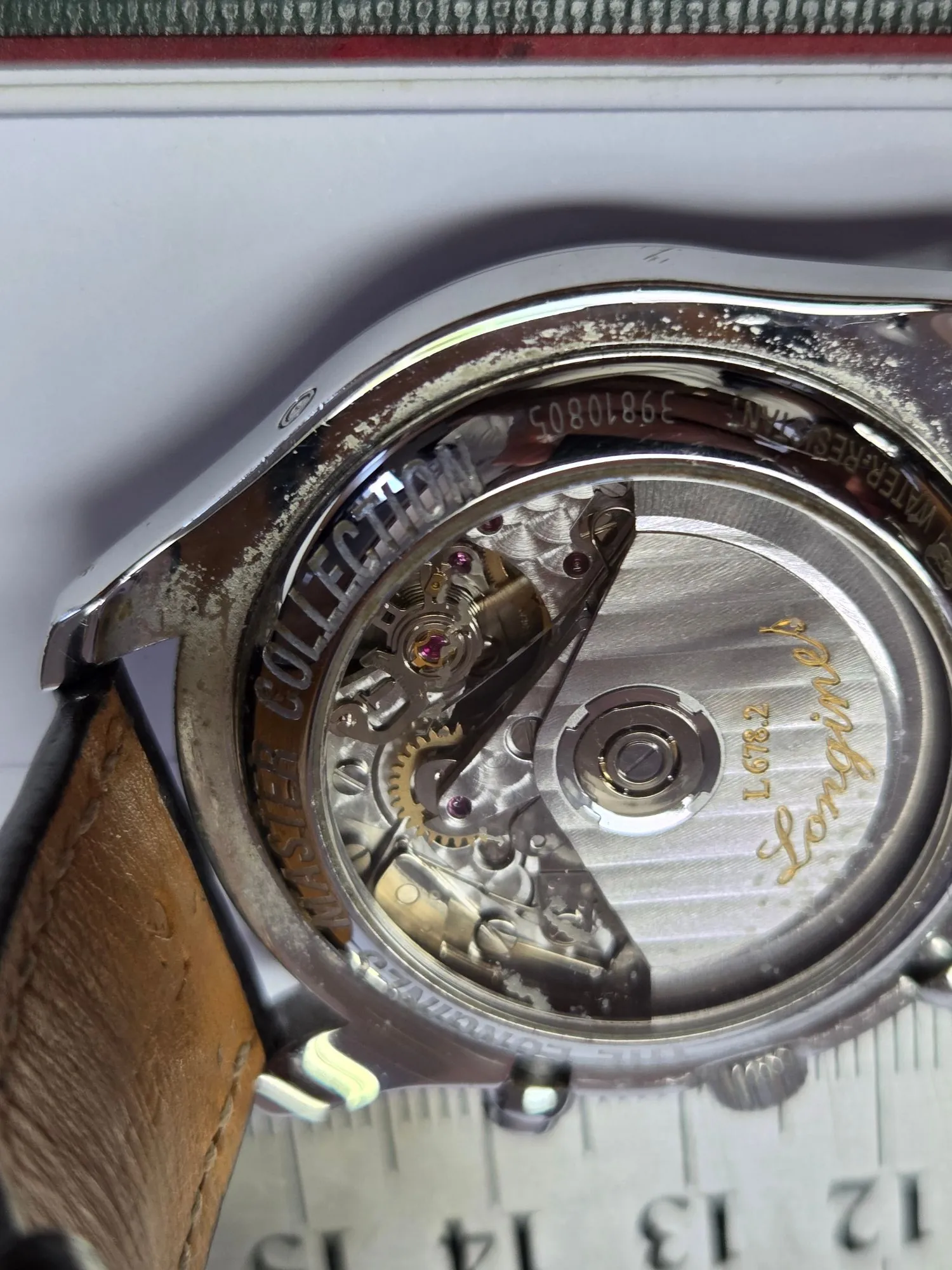 Zbliżenie na tył zegarka marki Longines z przezroczystym dekielkiem, przez który widoczny jest mechanizm z kołami zębatymi i wahadłem. Na metalowej obudowie widnieją napisy: 'MASTER COLLECTION', 'L678.2', 'Longines' oraz numer seryjny '39810805' i napis 'WATER RESIST'. Zegarek ma brązowy skórzany pasek i leży na białym tle obok linijki z widocznymi liczbami 12, 13, 14.