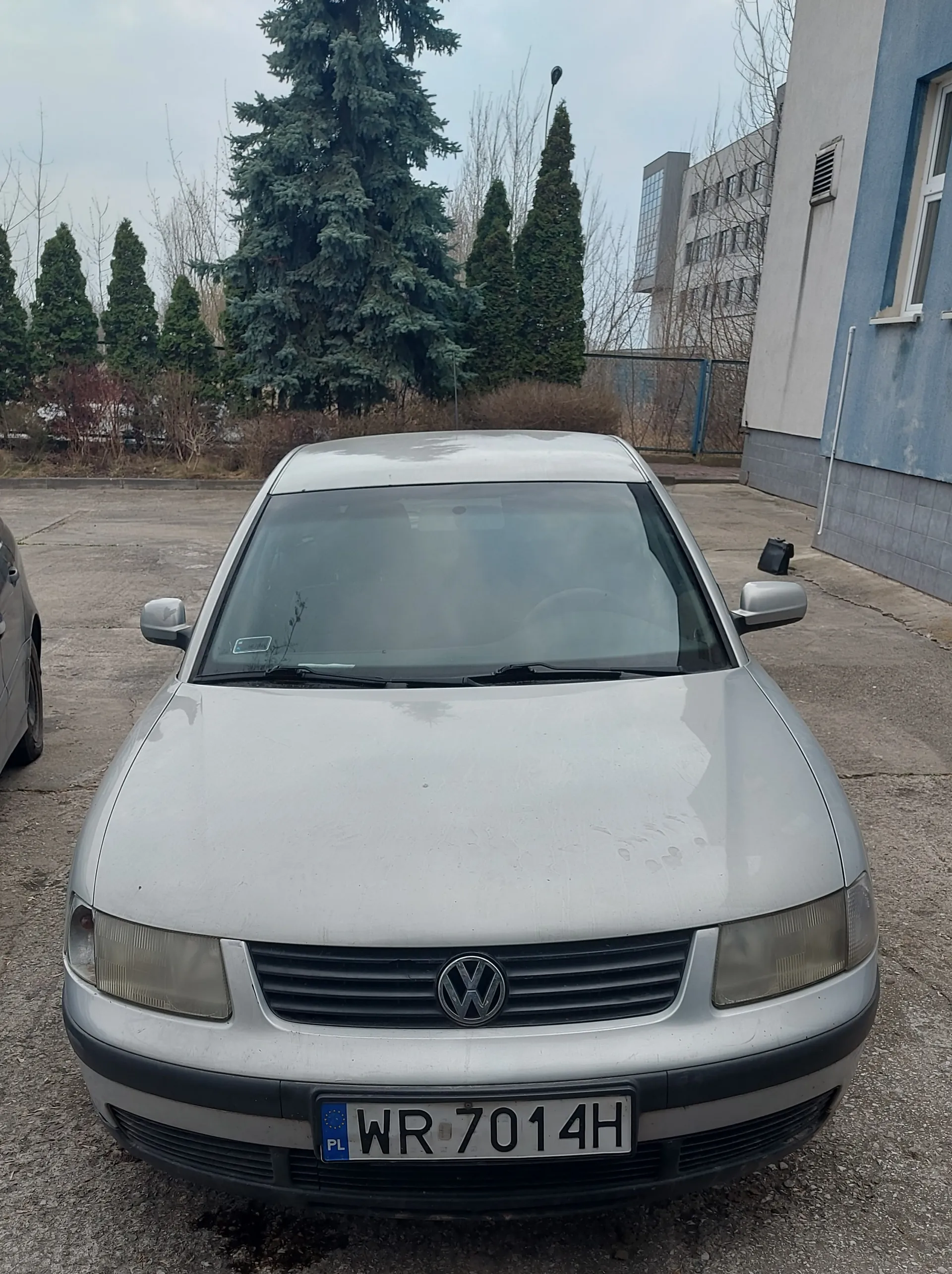 Srebrny samochód marki Volkswagen z tablicą rejestracyjną WR 7014H (PL) zaparkowany na podwórku. W tle widoczne są drzewa iglaste, budynek z niebiesko-szarymi ścianami, ogrodzenie oraz fragment innego pojazdu po lewej stronie. Niebo jest zachmurzone.
