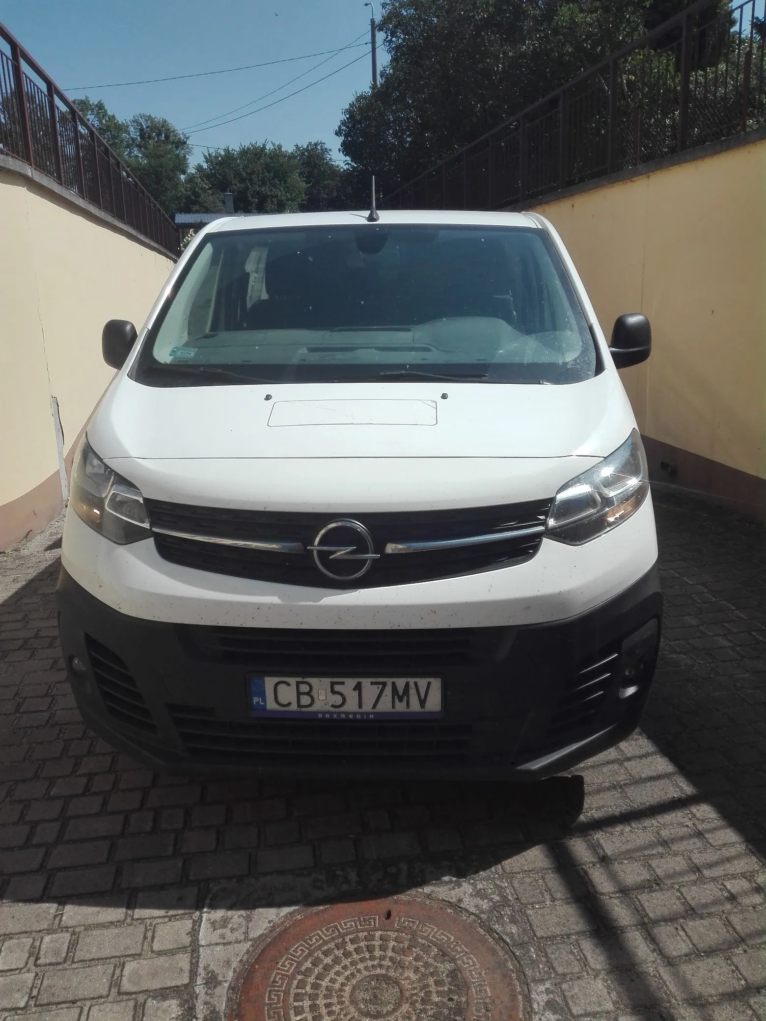 Zdjęcie przodu białego vana marki Opel zaparkowanego na kostce brukowej. Pojazd ma czarny zderzak i polską tablicę rejestracyjną o numerze CB 517MV. W tle widoczne są żółte mury, barierki oraz zielone drzewa pod jasnym niebem.