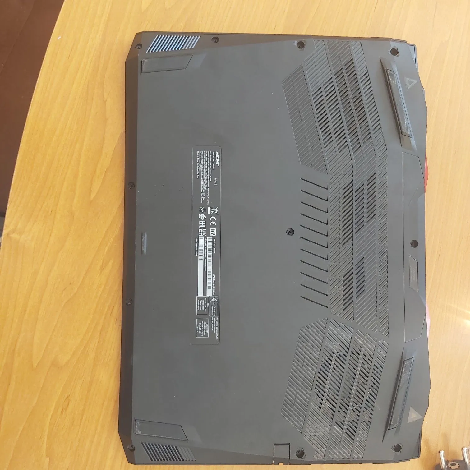 Zdjęcie przedstawiające spód czarnego laptopa marki Acer, leżącego na drewnianym blacie. Na obudowie widoczne są kratki wentylacyjne, gumowe stopki oraz etykieta z informacjami technicznymi, logo Acer, oznaczeniami CE, FCC oraz numerem seryjnym. Tekst na etykiecie jest zorientowany pionowo względem krawędzi laptopa.