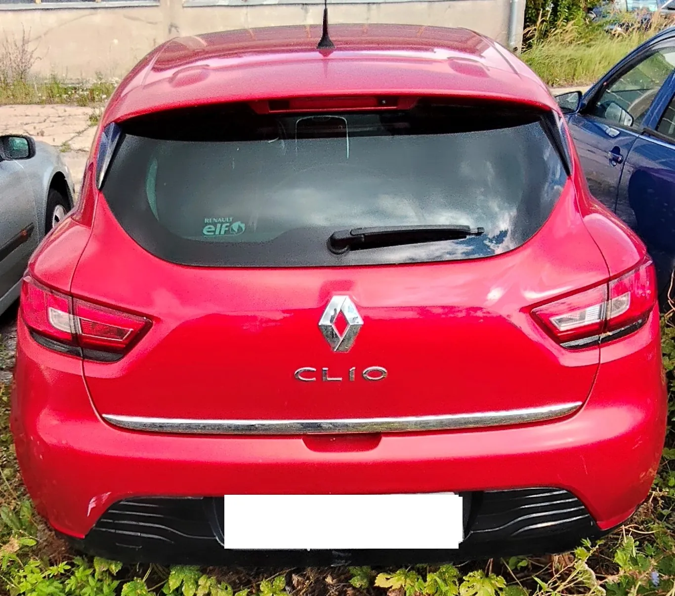 Tylny widok czerwonego samochodu marki Renault Clio. Na klapie bagażnika znajduje się srebrne logo Renault oraz napis 'CLIO'. Tablica rejestracyjna jest zakryta białym prostokątem. Po lewej stronie widoczny jest fragment srebrnego samochodu, a po prawej fragment niebieskiego samochodu.