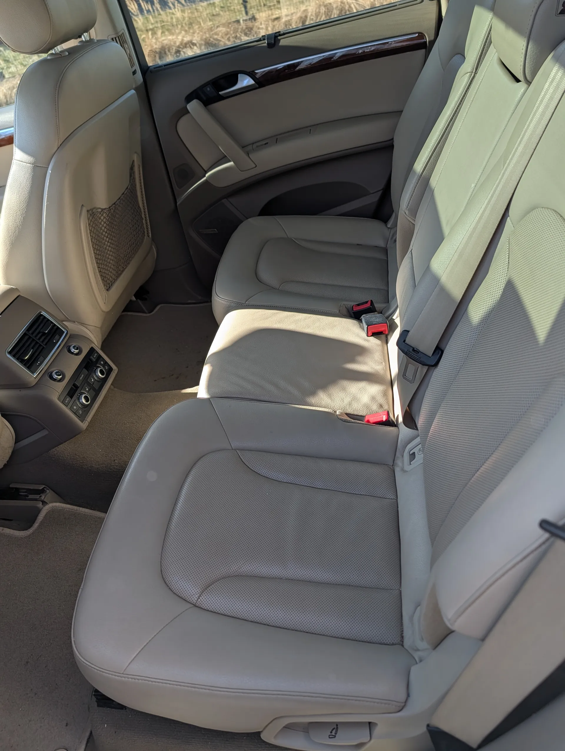 Wnętrze samochodu z beżowymi skórzany siedzeniami, widok na przednie siedzenia z pasami bezpieczeństwa, centralną konsolę z wentylacją i przyciskami sterowania, panel drzwiowy z uchwytem i sterowaniem okien, a przez okno widoczna trawa na zewnątrz.