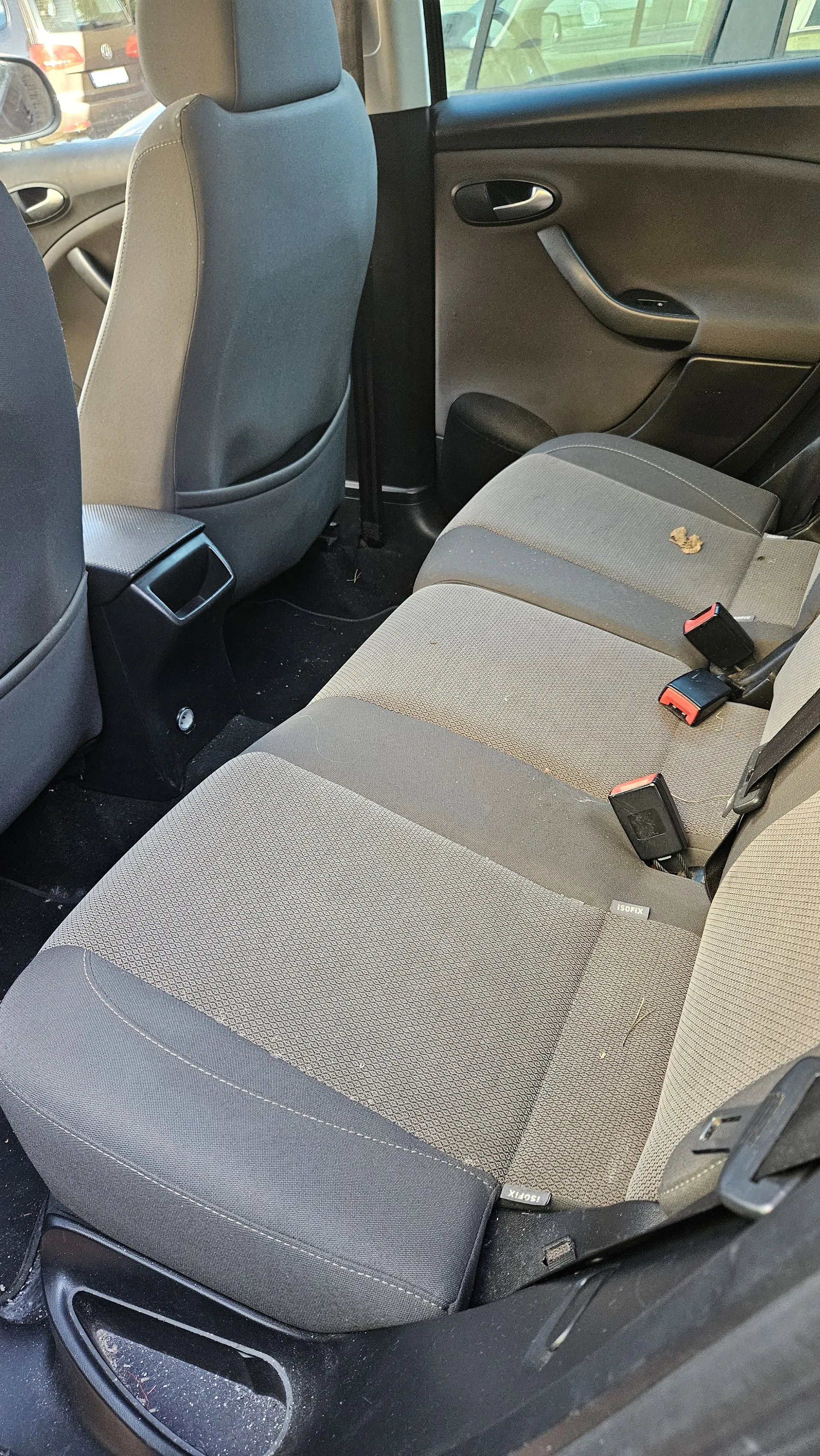 Wnętrze samochodu z widokiem na tylne siedzenia. Siedzenia w kolorze szaro-beżowym z widocznymi elementami pasów bezpieczeństwa o czerwono-czarnych zapięciach. Na siedzeniu znajduje się mały brązowy plamka lub śmieci. Widoczne są fragmenty przednich siedzeń, drzwi z uchwytem i oknem, a na zewnątrz częściowo inny pojazd. Na siedzeniu widnieje napis ISOFIX.