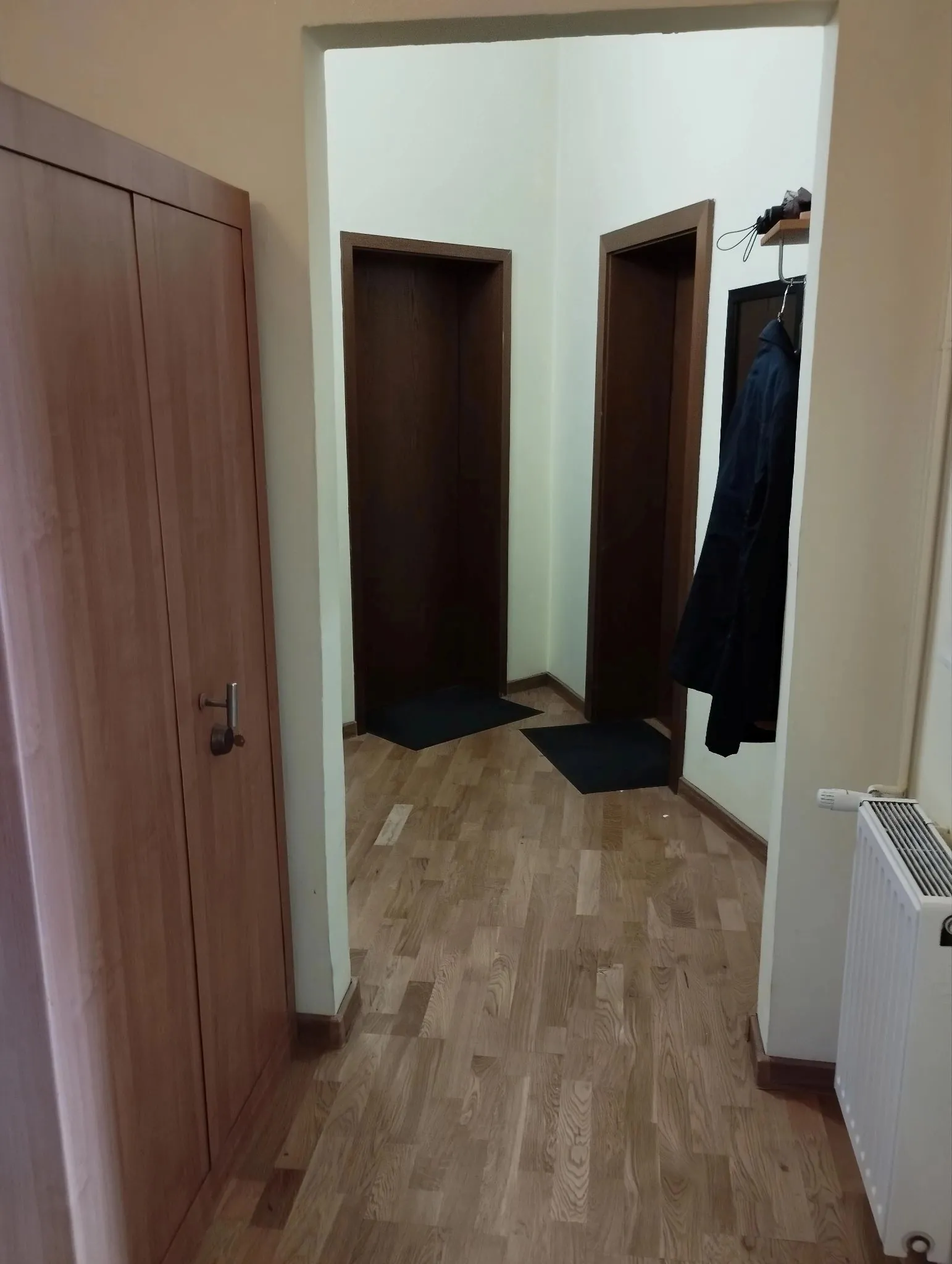 Widok na przedpokój z jasnymi ścianami i drewnianą podłogą. Po lewej stronie znajduje się duża, drewniana szafa z dwiema drzwiami. W głębi widoczne są dwa otwarte przejścia do innych pomieszczeń, przed którymi leżą ciemne wycieraczki. Po prawej stronie wisi czarny płaszcz na wieszaku, a poniżej znajduje się biały grzejnik panelowy.