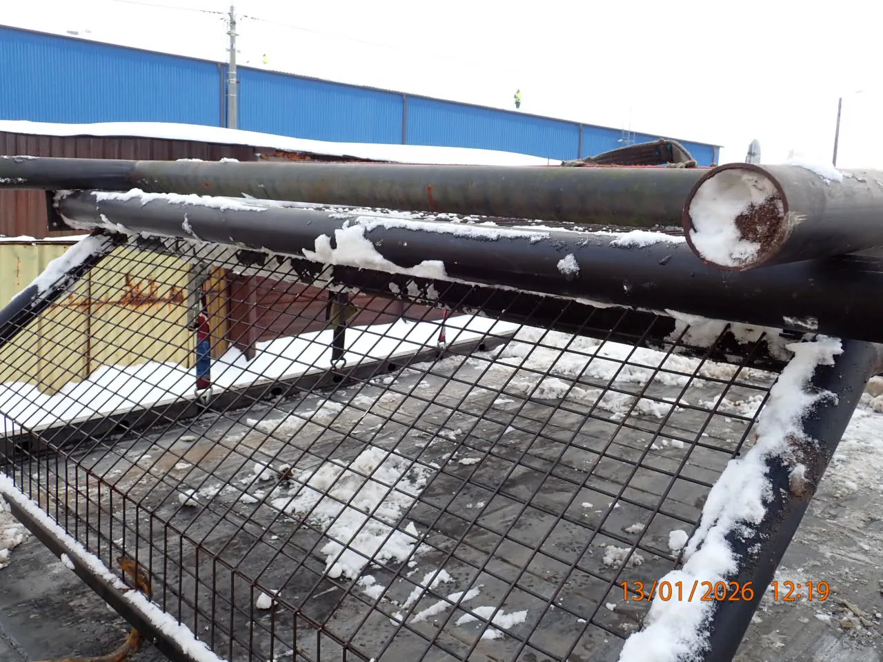 Przemysłowy obiekt z metalowymi rurami pokrytymi śniegiem, siatką stalową i budynkiem o niebieskiej elewacji w tle. Na pierwszym planie widoczne są duże, poziome rury z resztkami śniegu, a pod nimi metalowa siatka z zasnieżonymi fragmentami. W tle budynek z niebieską blachą falistą i dachem pokrytym śniegiem. W prawym dolnym rogu obrazu znajduje się pomarańczowy znacznik czasu: 13/01/2026 12:19. Podłoże pokryte jest śniegiem i lodem.