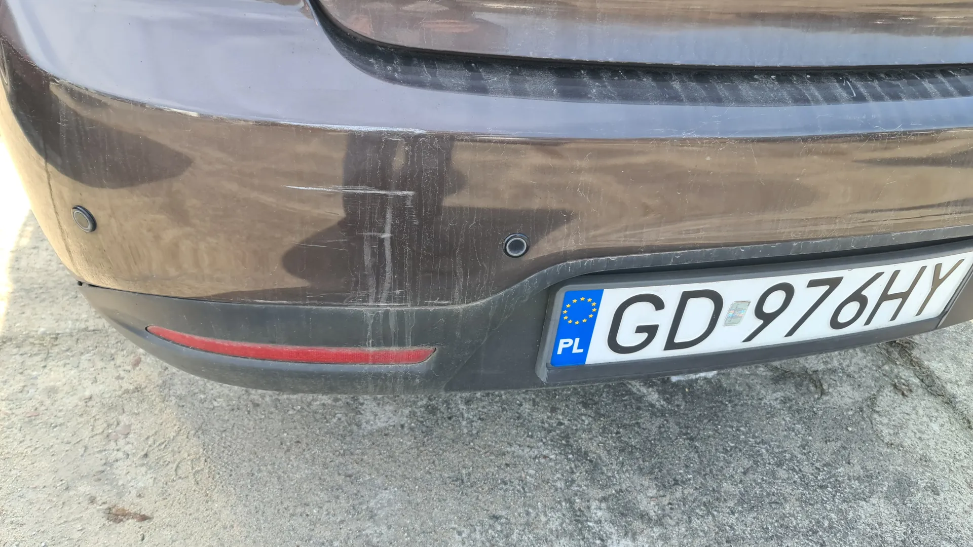 Zbliżenie na tylną część brązowego samochodu z widoczną tablicą rejestracyjną. Tablica ma biały tło z czarnym napisem GD 976HY, niebieską częścią z napisem PL i żółtymi gwiazdkami Unii Europejskiej. Na zderzaku widoczne są dwa czarne czujniki parkowania oraz czerwony odblask. Samochód ma ślady zabrudzeń i zadrapań na karoserii. Tło stanowi szary asfalt.