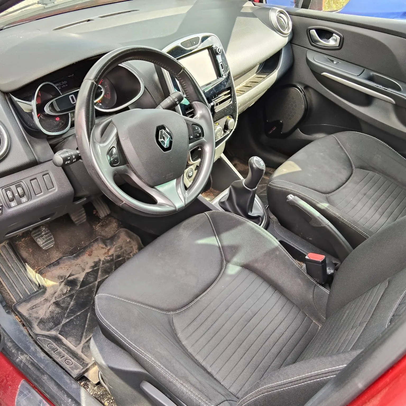 Wnętrze samochodu marki Renault Clio widziane z perspektywy otwartych drzwi pasażera. Widoczna jest czarna kierownica z logo Renault, deska rozdzielcza z ekranem multimedialnym, liczniky oraz dwa przednie fotele obite czarnym materiałem z przeszyciami. Na podłodze znajduje się gumowa mata z napisem 'CLIO'.