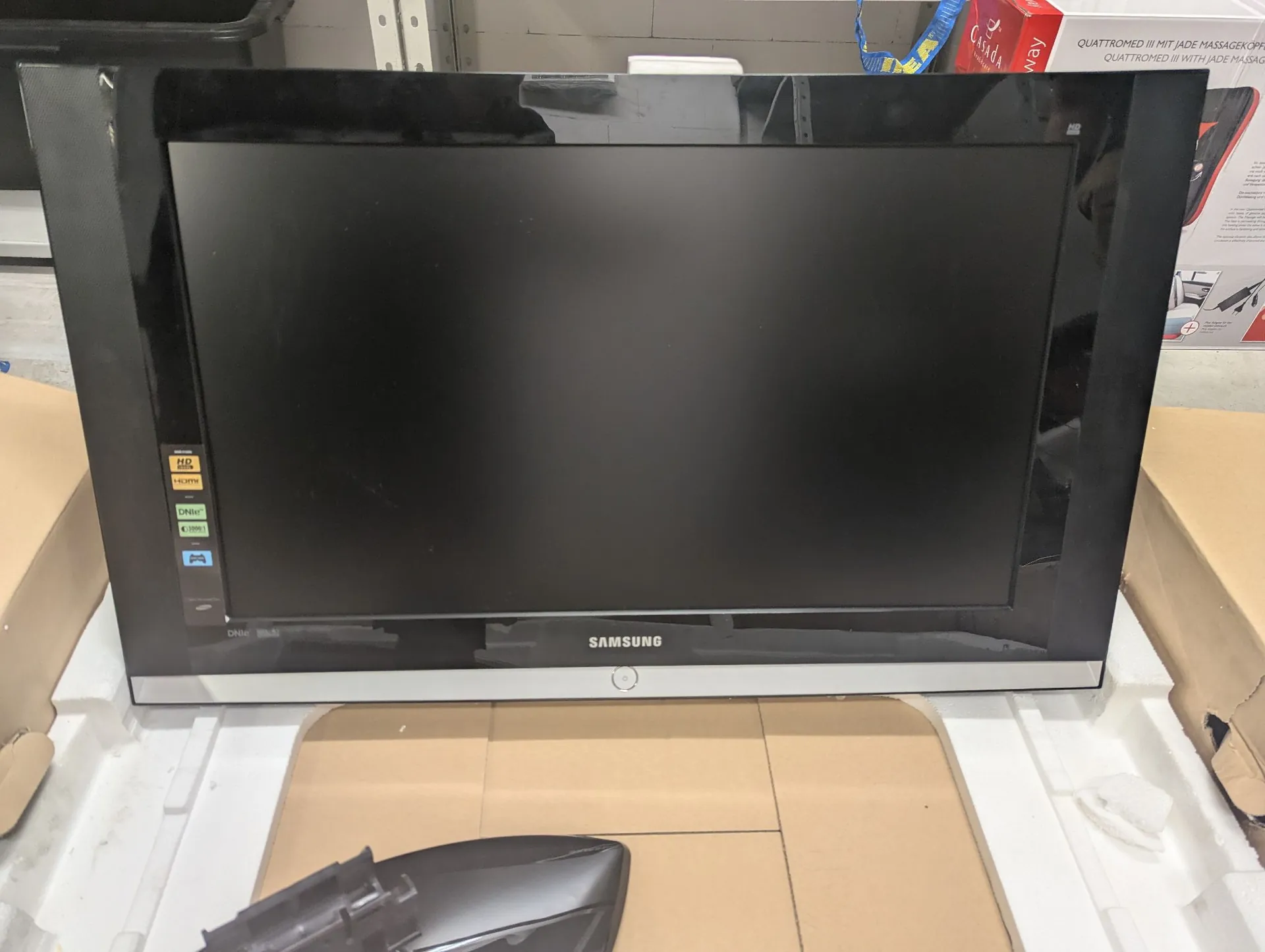 Czarny telewizor marki Samsung z srebrną ramką na dole, umieszczony w białym styropianowym opakowaniu transportowym. Na ekranie po lewej stronie widoczne są małe kolorowe naklejki z oznaczeniami technicznymi. Na dolnej srebrnej krawędzi znajduje się biały napis 'SAMSUNG'.