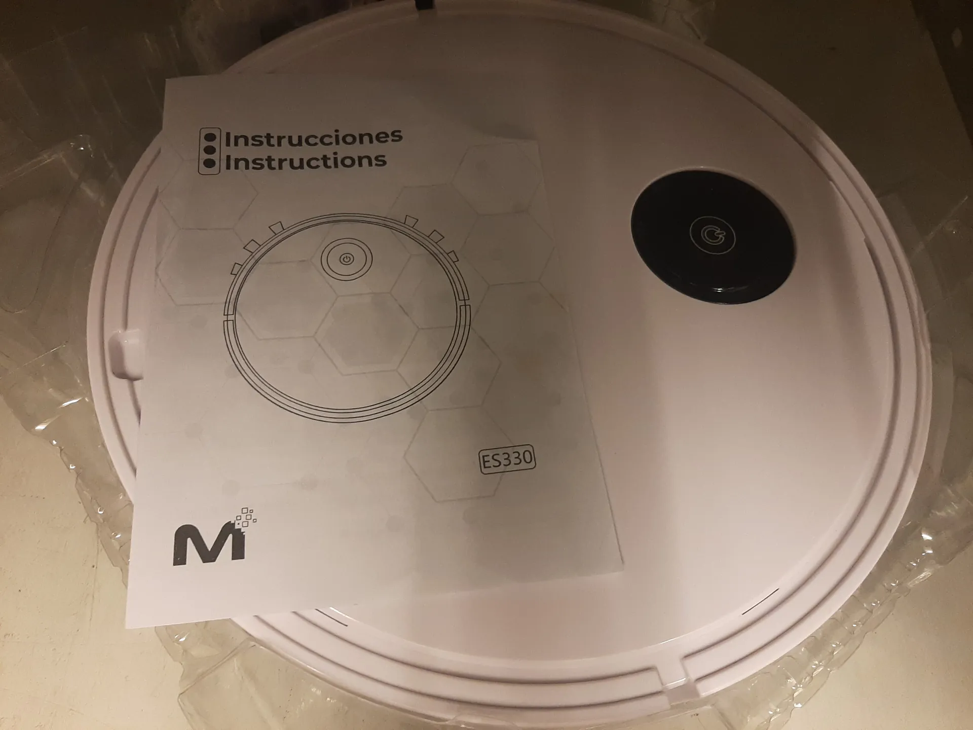 Biały, okrągły urządzenie z czarnym okrągłym przyciskiem z symbolem zasilania. Na urządzeniu leży arkusz z napisem "Instrucciones Instructions", schematem urządzenia, oznaczeniem "ES330" oraz logo z literą "M" i kropkami. Urządzenie znajduje się na przezroczystej folii. Tekst na arkuszu jest czytelny i ułożony poziomo.