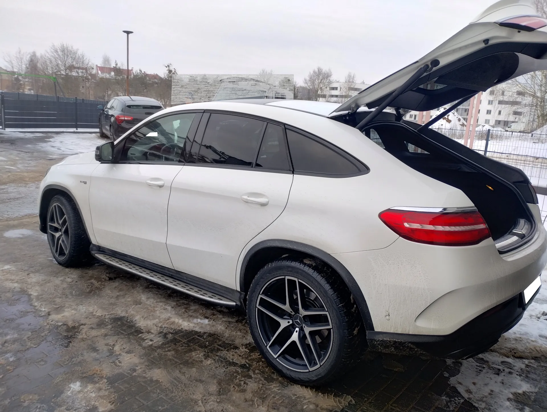 Biały SUV z otwartym bagażnikiem zaparkowany na mokrym, błotnistym terenie z resztkami śniegu. W tle czarny samochód, ogrodzenie, budynki i bezlistne drzewa. Niebo zachmurzone.