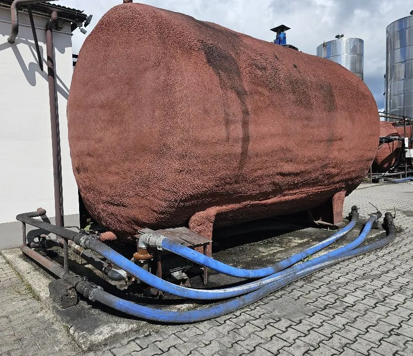 Duży, poziomy zbiornik o cylindrycznym kształcie z półkolistymi końcami, pokryty chropowatą, brązowo-rdzawą izolacją. Zbiornik spoczywa na metalowych podporach na betonowym podłożu z kostki brukowej. Z dołu zbiornika wychodzą rury i trzy grube, niebieskie węże prowadzące w prawą stronę obrazu. W tle widoczne są białe ściany budynku oraz dwa wysokie, srebrne, metalowe silosy pod zachmurzonym niebem.