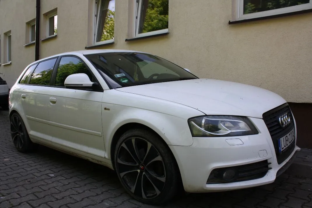 Biały samochód marki Audi w nadwoziu kombi, zaparkowany na kostce brukowej obok jasnej ściany budynku z oknami. Pojazd posiada ciemne, pięcioramienne felgi. Widoczna jest część tablicy rejestracyjnej z tekstem 'LUB 221'.