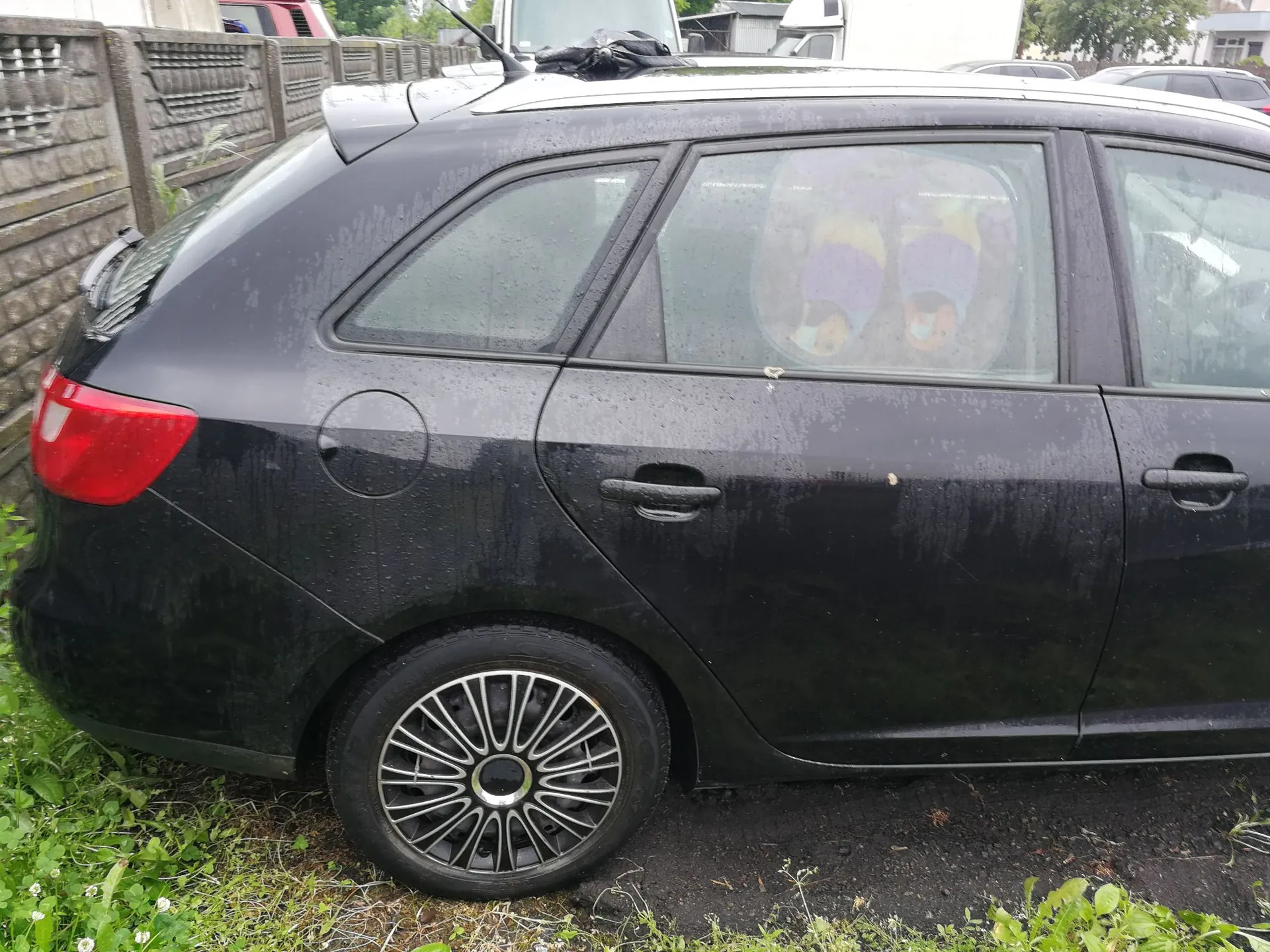 Boczny widok tylnej części czarnego samochodu typu kombi. Auto jest brudne, z widocznymi zaciekami i kroplami wody na lakierze. W tylnej szybie odbija się postać w fioletowych okularach. Samochód stoi na trawiastym poboczu obok betonowego ogrodzenia.