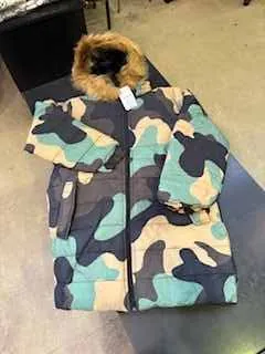 Kurtka w kamuflażu zielono-czarno-beżowym z futrzanym kołnierzem i białą etykietą na kołnierzu. Kurtka leży na powierzchni, na tle widoczne są ciemne i jasne obszary.