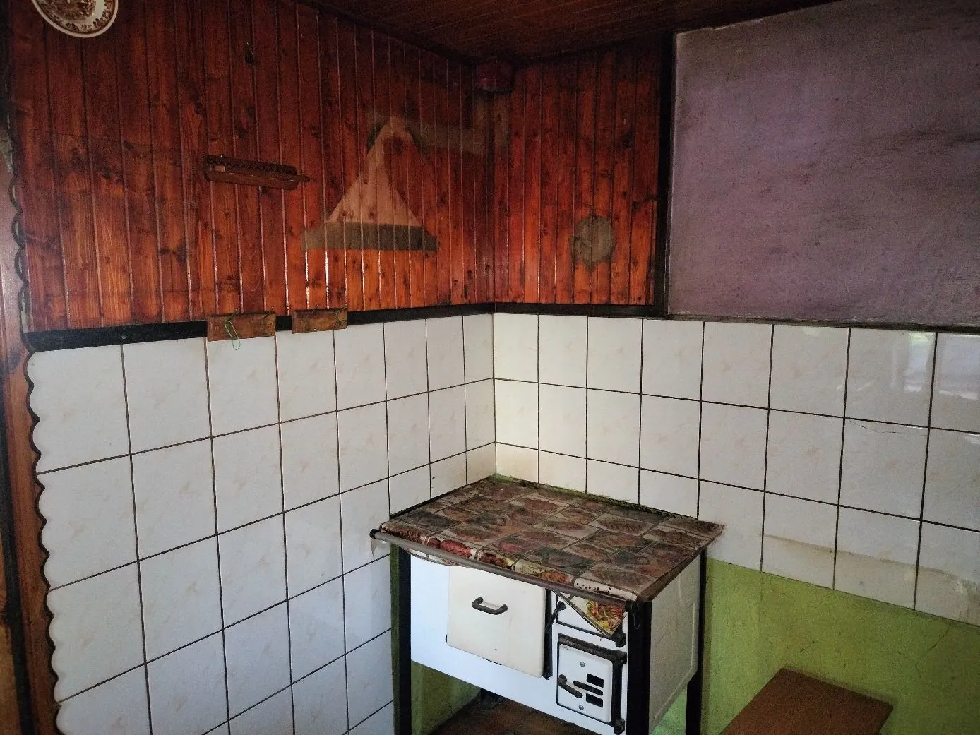 Wnętrze pomieszczenia z białymi kafelkami na dolnej części ścian i drewnianą boazerią na górze. W rogu stoi mały, biały piecyk lub kuchenka z blatem w kolorowe wzory. Po prawej stronie widoczna jest gładka, fioletowa ściana.