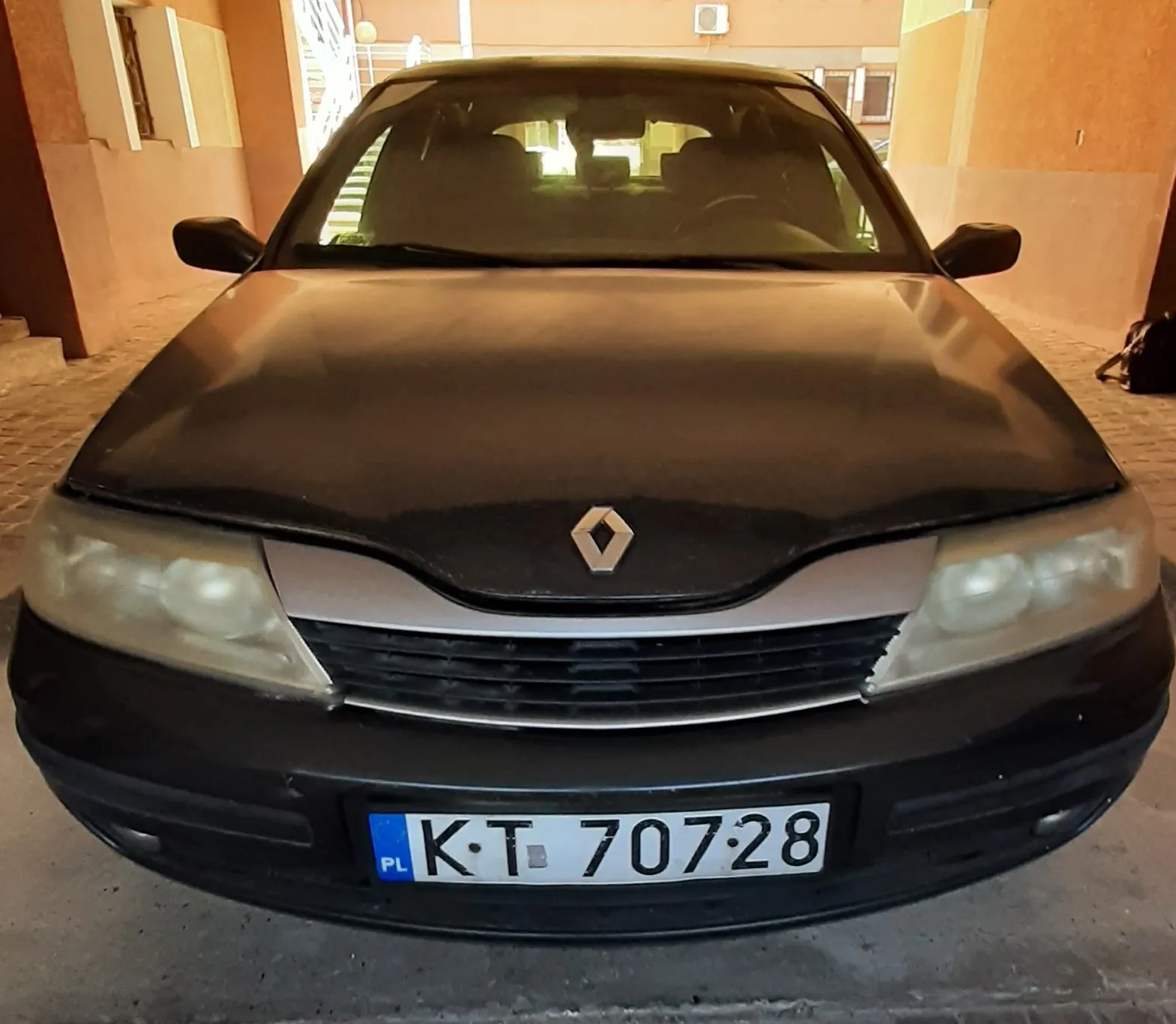 Czarny samochód marki Renault z przodu, z widocznym logo na masce. Tablica rejestracyjna z polskim oznaczeniem PL i numerem KT 70728. Samochód zaparkowany w podwórku lub garażu, na tle budynku z pomarańczowymi ścianami i schodami. Po prawej stronie widoczna czarna torba na podłodze.