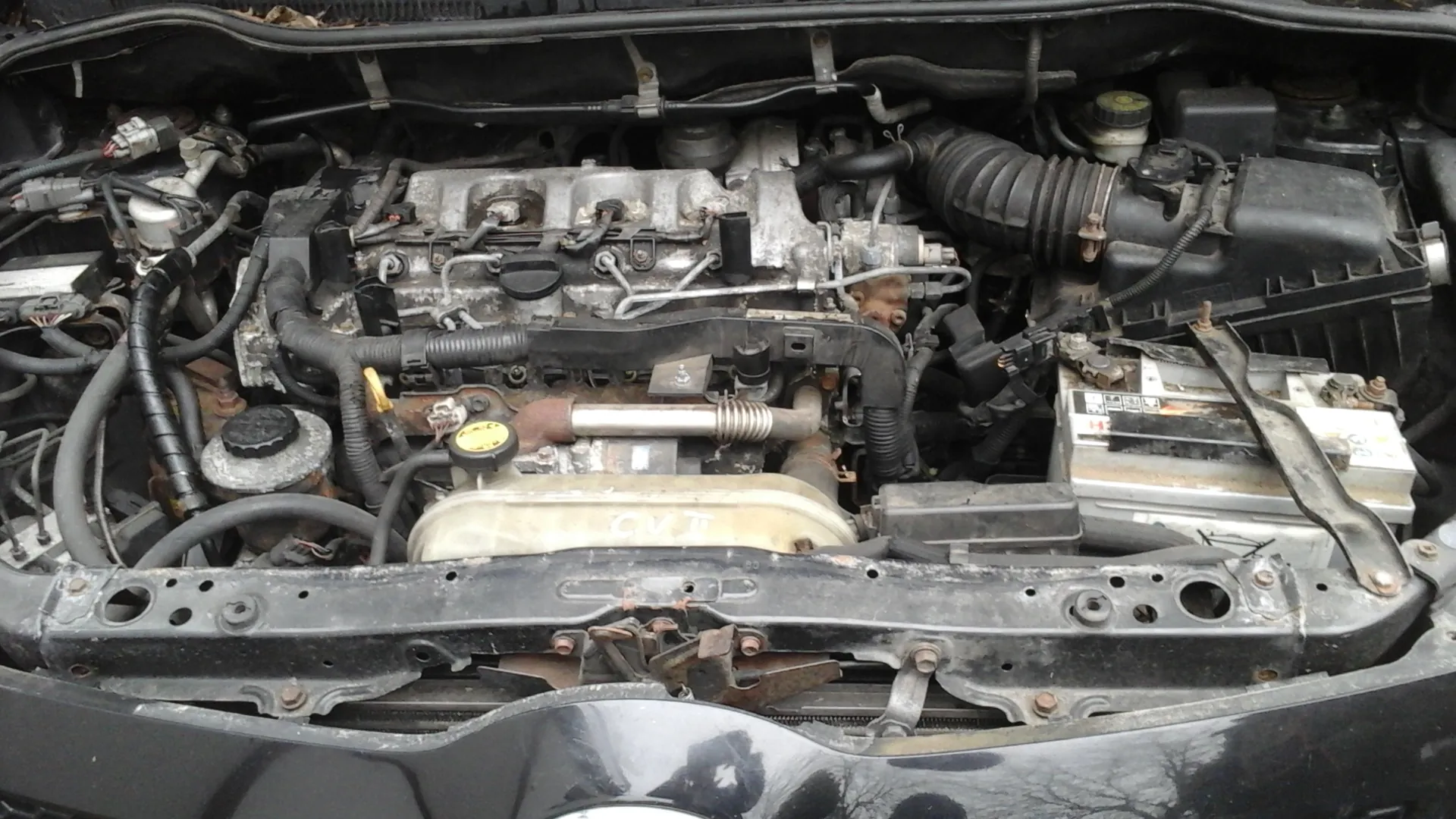 Zdjęcie przedstawia otwarty komorę silnika samochodu. Widoczne są elementy takie jak przewody, węże, akumulator z częściowo czytelnymi oznaczeniami, pokrywa silnika z napisem CVT oraz zardzewiałe i zabrudzone części mechaniczne. W dolnej części obrazu znajduje się czarna karoseria pojazdu z widocznymi śladami korozji i uszkodzeń.